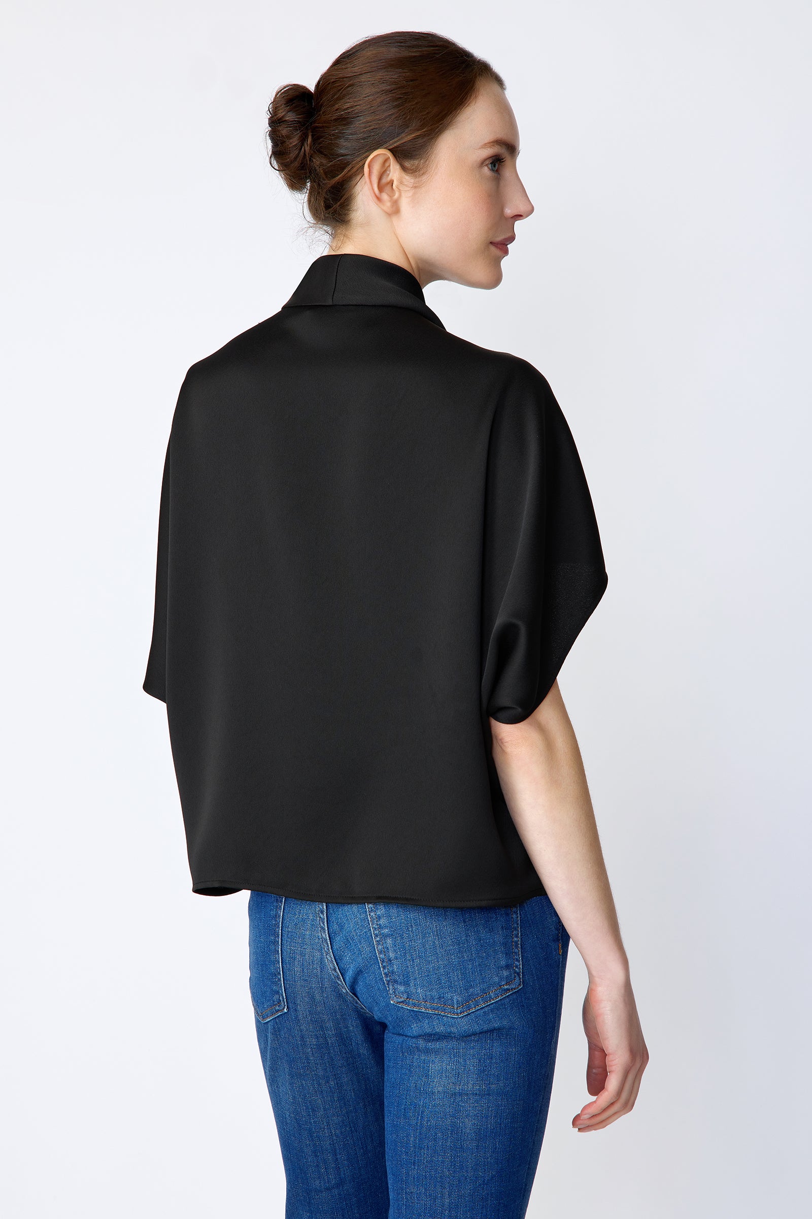 Woman wearing Kal Rieman Angelica Cape Top in Black and blue jeans on a white background, arm on the side back view