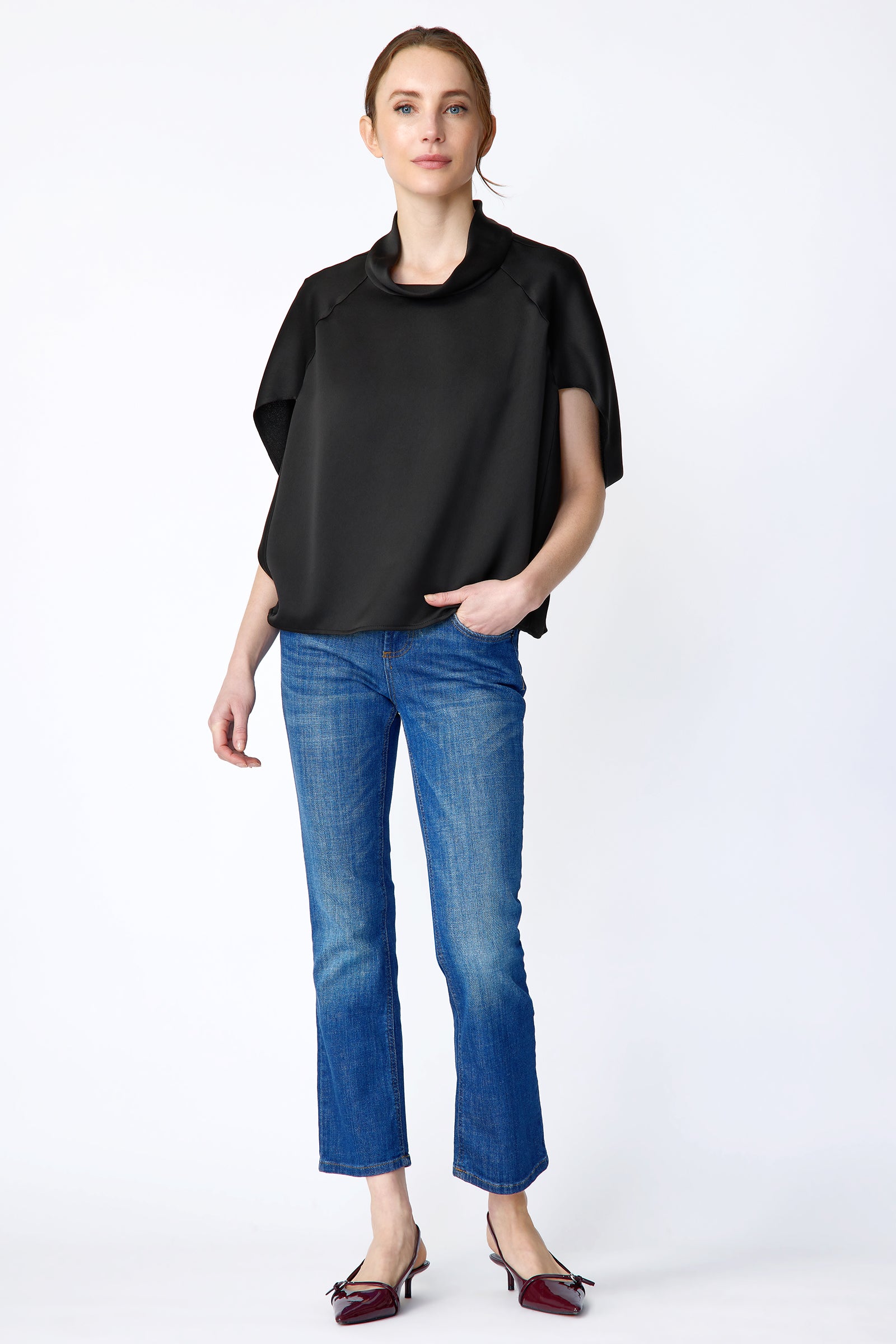 Woman wearing Kal Rieman Angelica Cape Top in Black and blue jeans on a white background, hand in pant pockets, arm on side