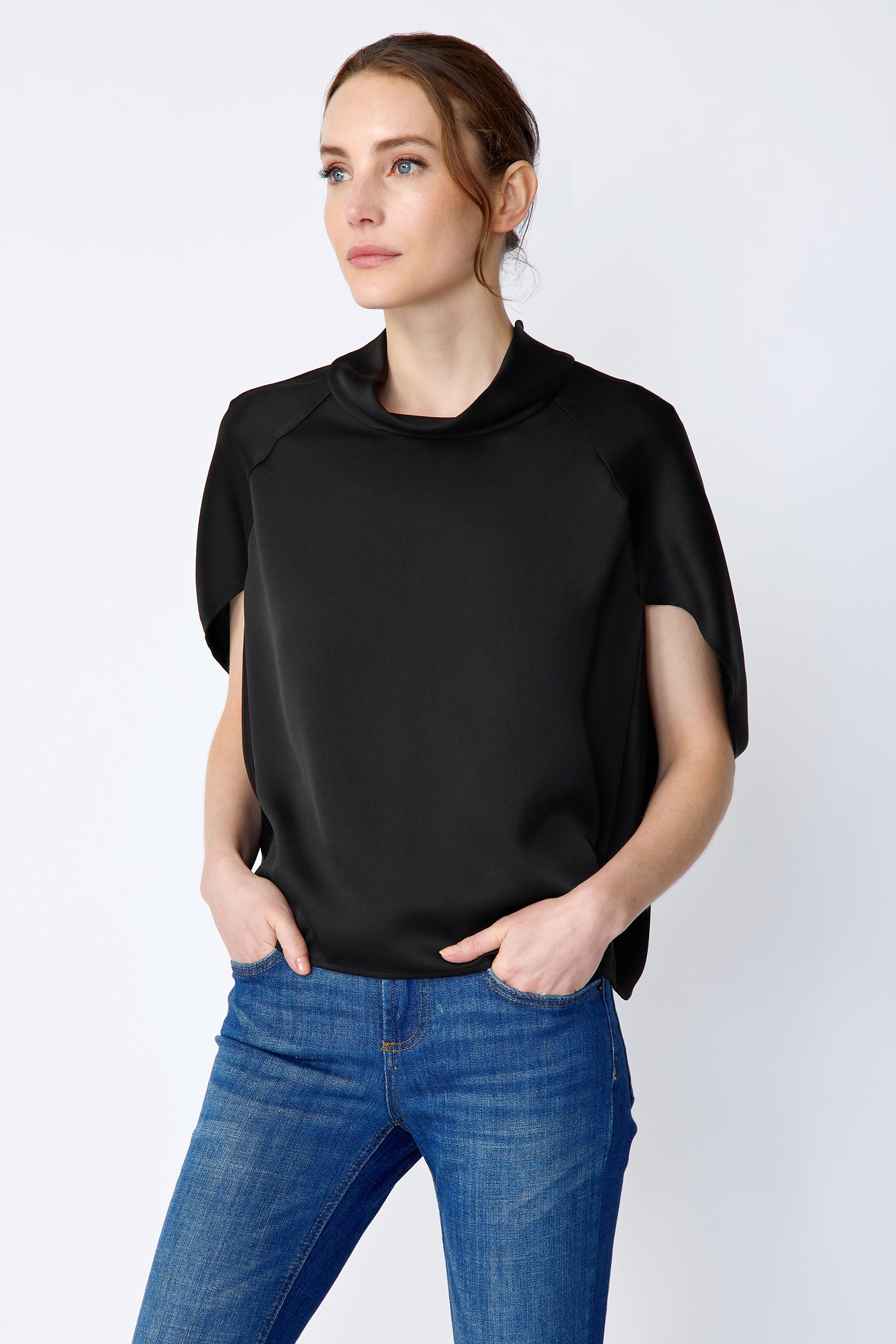 Woman wearing Kal Rieman Angelica Cape Top in Black and blue jeans on a white background, hands in pant pockets, looking side ways