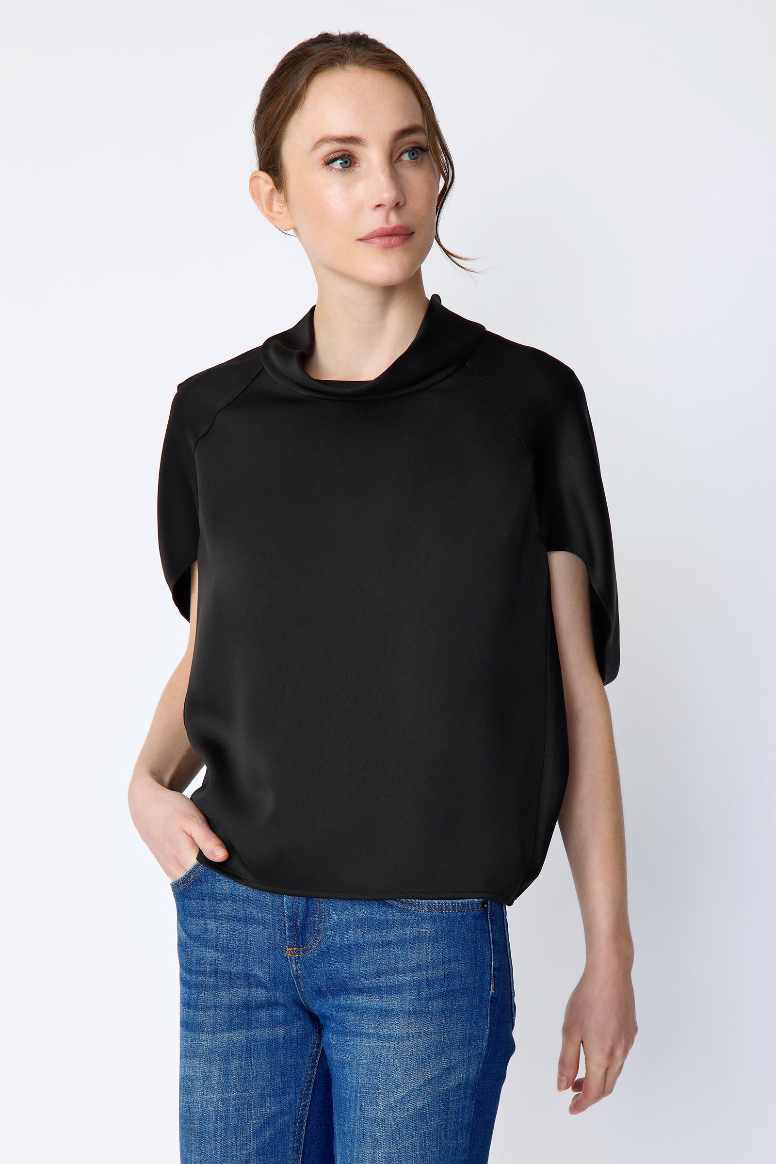 Woman wearing Kal Rieman Angelica Cape Top in Black and blue jeans on a white background, hand in pant pocket, arm on the side looking side way
