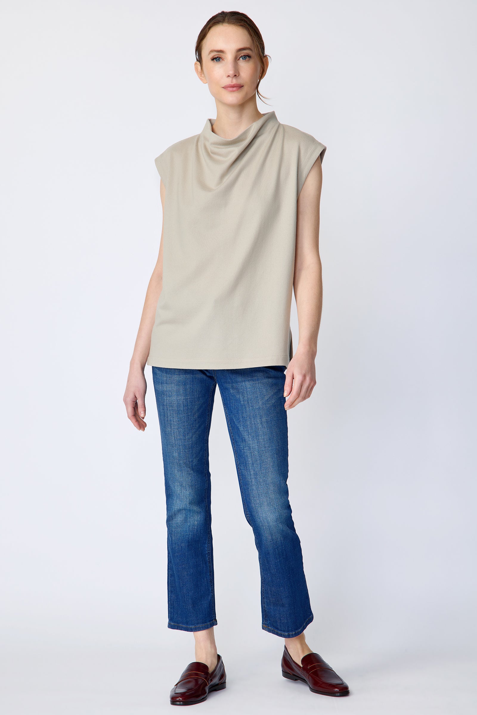 Woman wearing Kal Rieman Arla Drape Top in Mink Felted Jersey and blue jeans and brown shoes on a white background, arms on the side