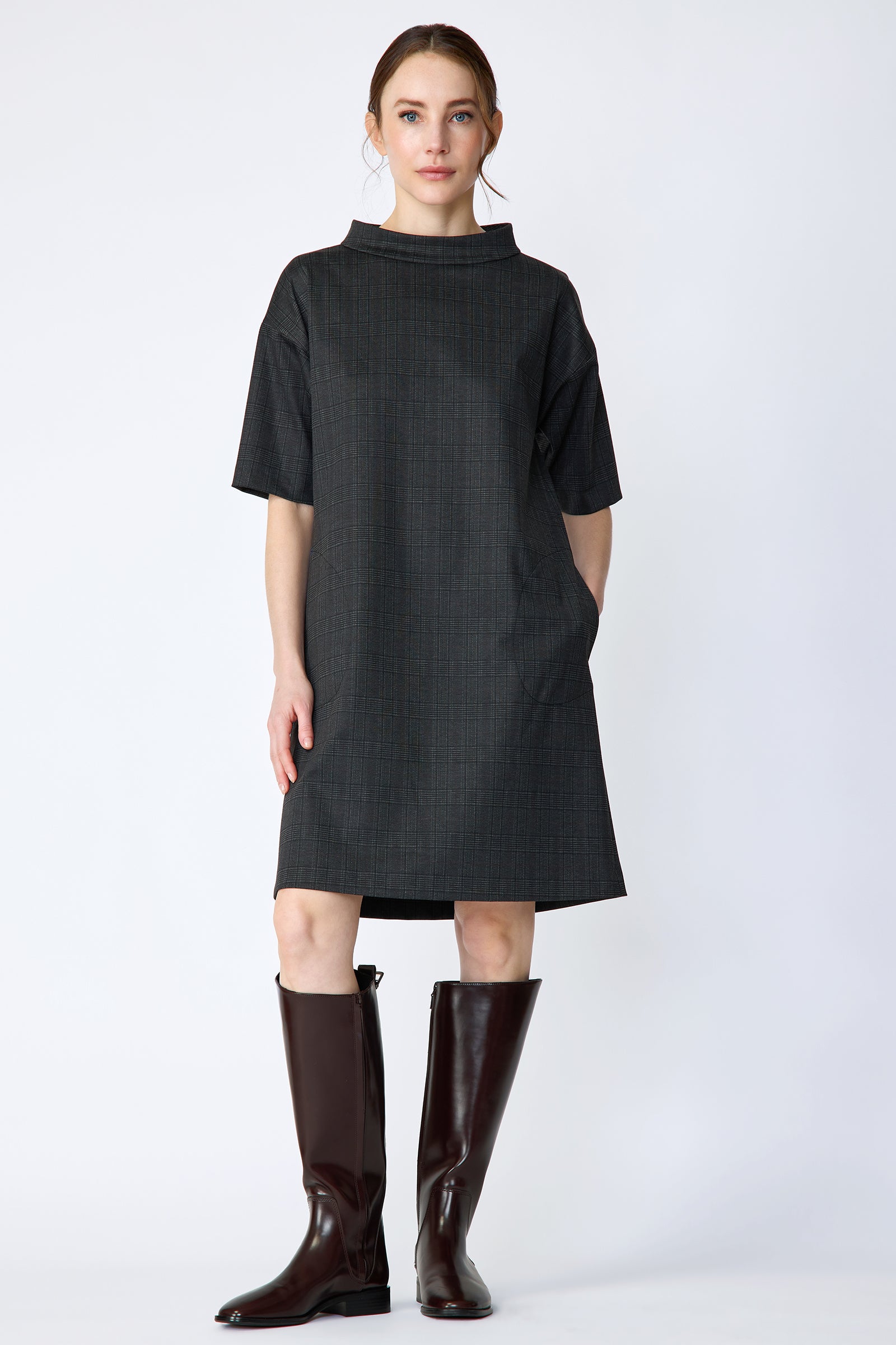 Woman wearing Kal Rieman Avery shift dress in black glen plaid and brown knee-high boots on a white background with one arm in pocket