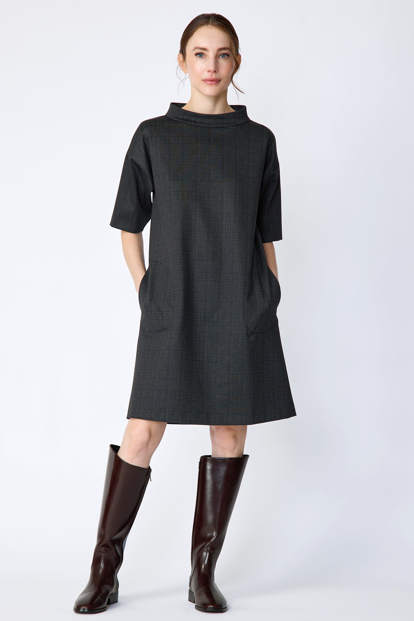 Woman wearing Kal Rieman Avery shift dress in black glen plaid and brown knee-high boots on a white background with arms in pockets