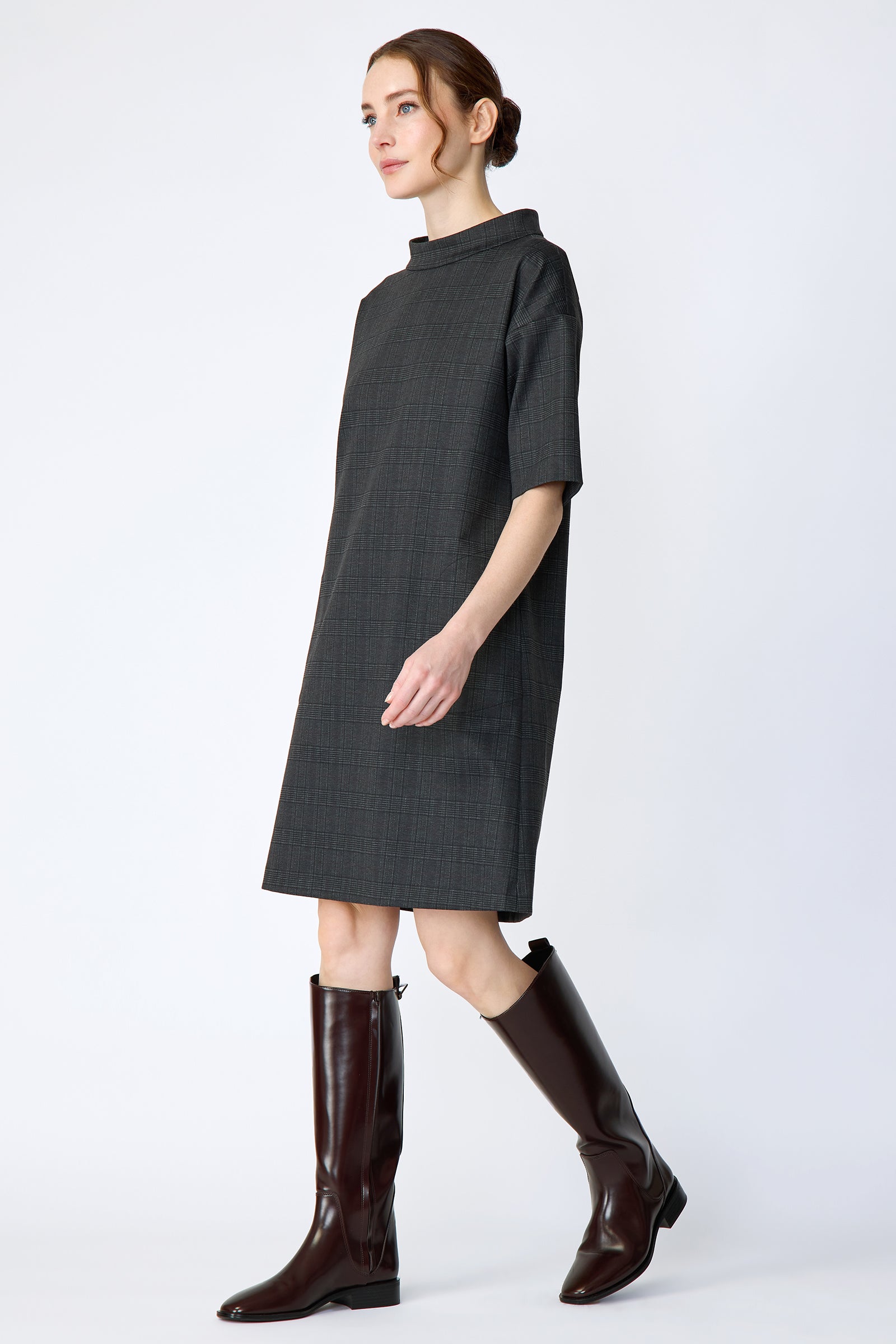 Woman wearing Kal Rieman Avery shift dress in black glen plaid and brown knee-high boots on a white background side view walking