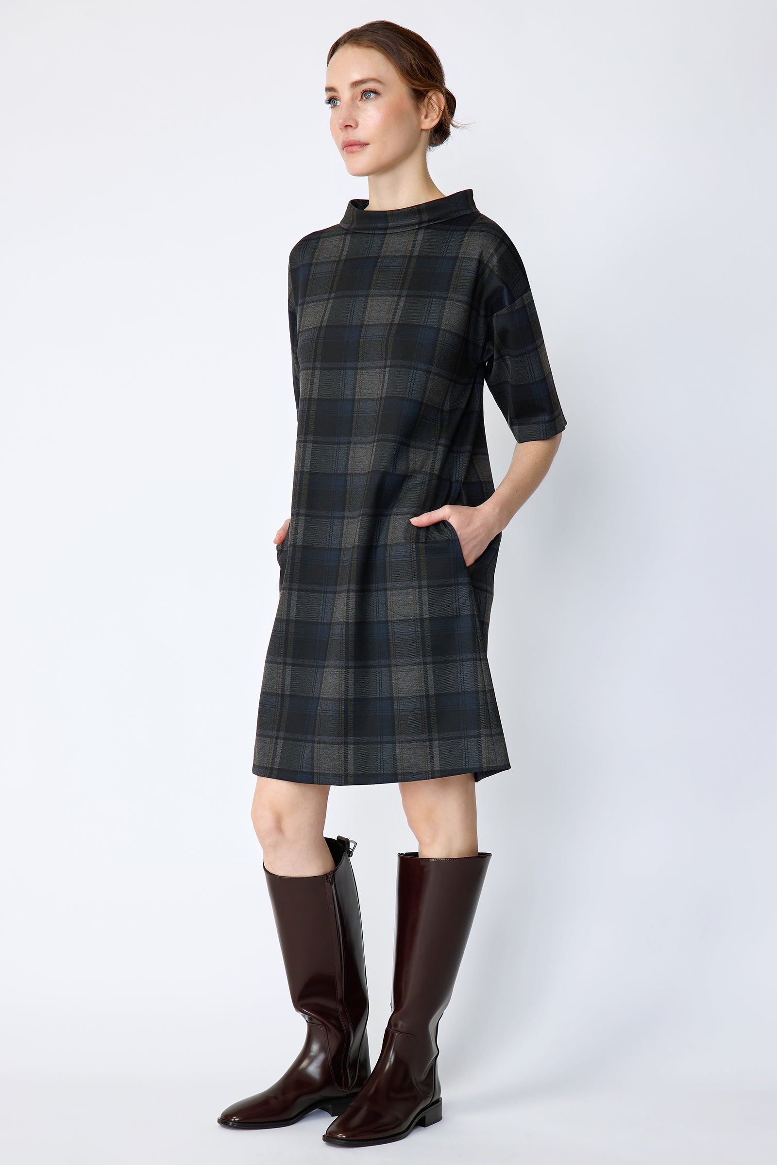 Woman wearing Kal Rieman Avery Shift Dress in Novelty Plaid with knee-high boots on a white background arms in pockets, side view