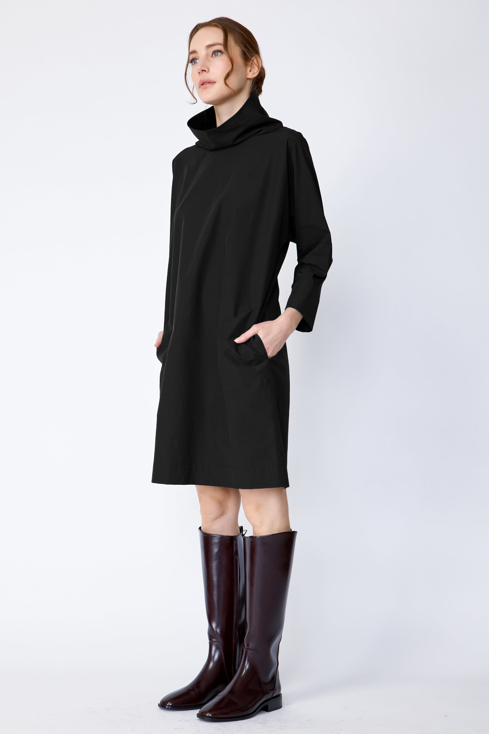 Woman wearing Kal Rieman Carla black dress and brown boots on a white background side view hands in pockets