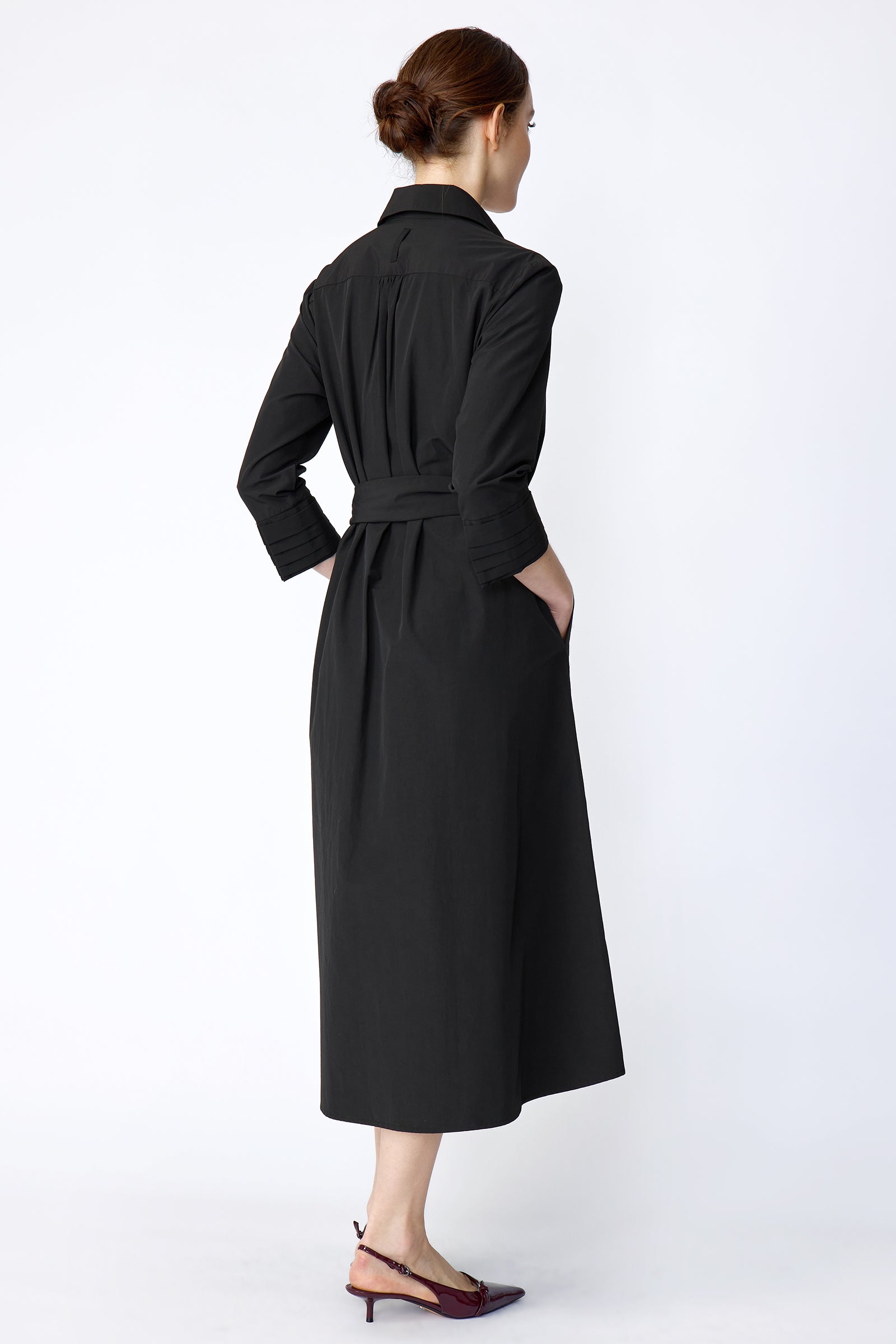 Woman wearing Kal Rieman Carolina Dress in Black Italian Broadcloth with burgundy shoes, against a white background, hands in pockets, back view