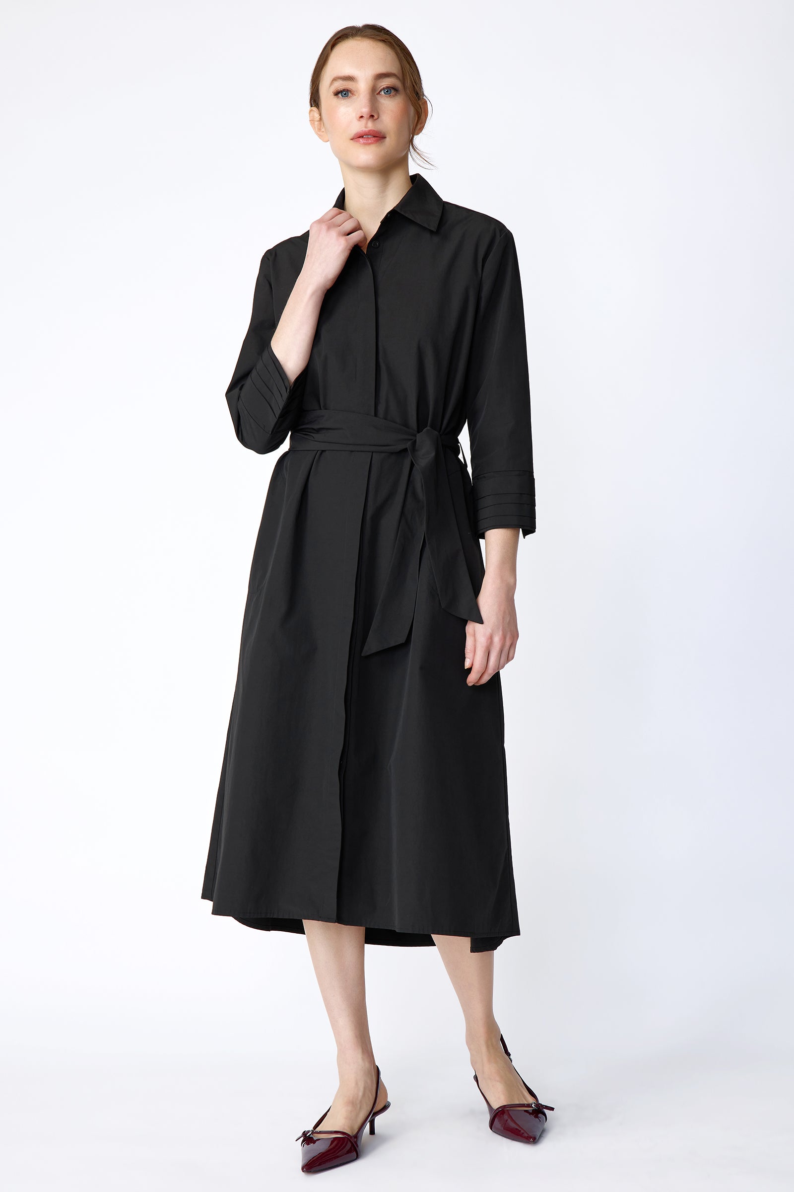 Woman wearing Kal Rieman Carolina Dress in Black Italian Broadcloth with burgundy shoes, against a white background, hand holding collar, arm on the side, full front view