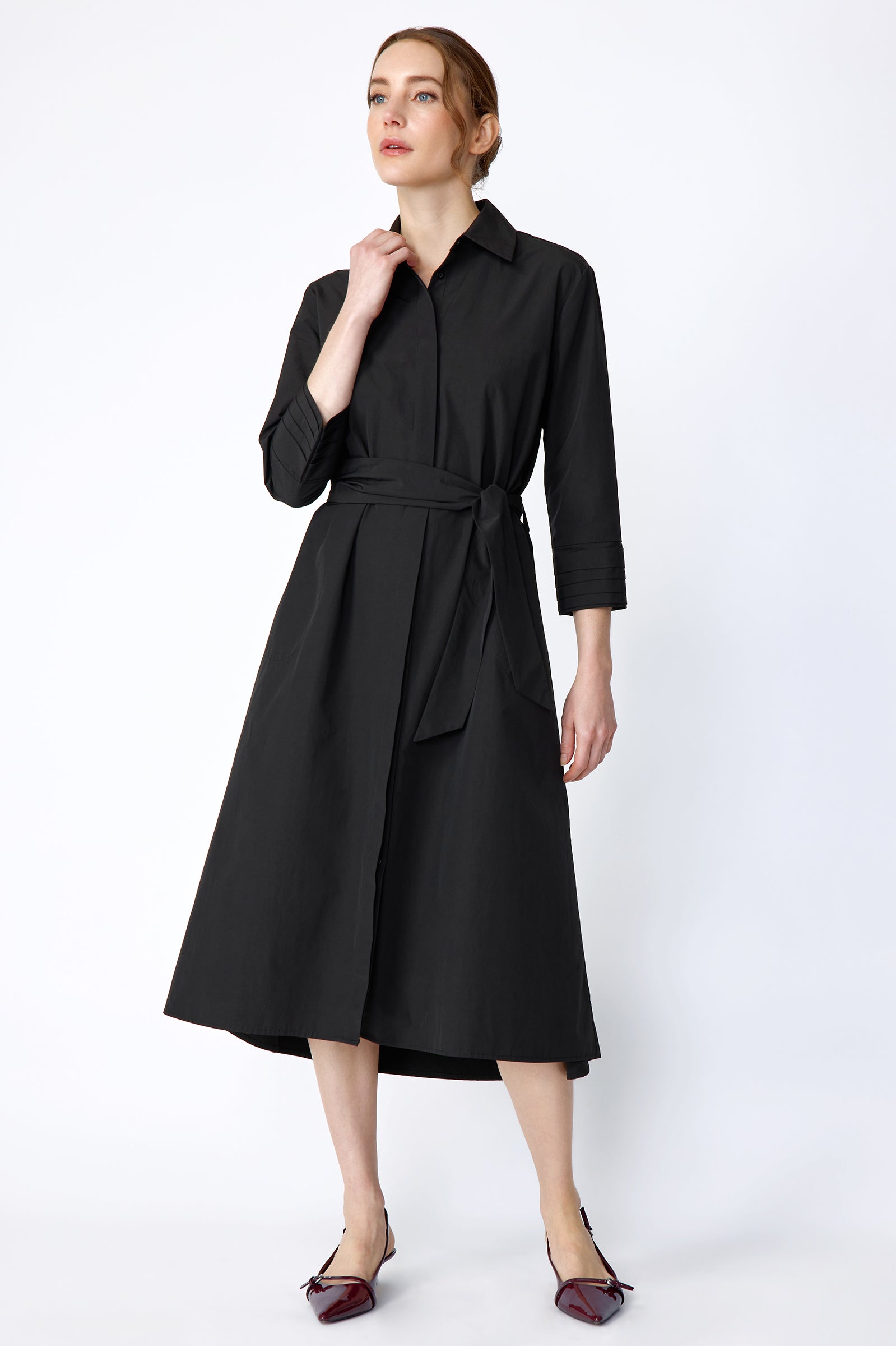 Woman wearing Kal Rieman Carolina Dress in Black Italian Broadcloth with burgundy shoes, against a white background, hand holding collar, arm on the side, looking sideways, full front view