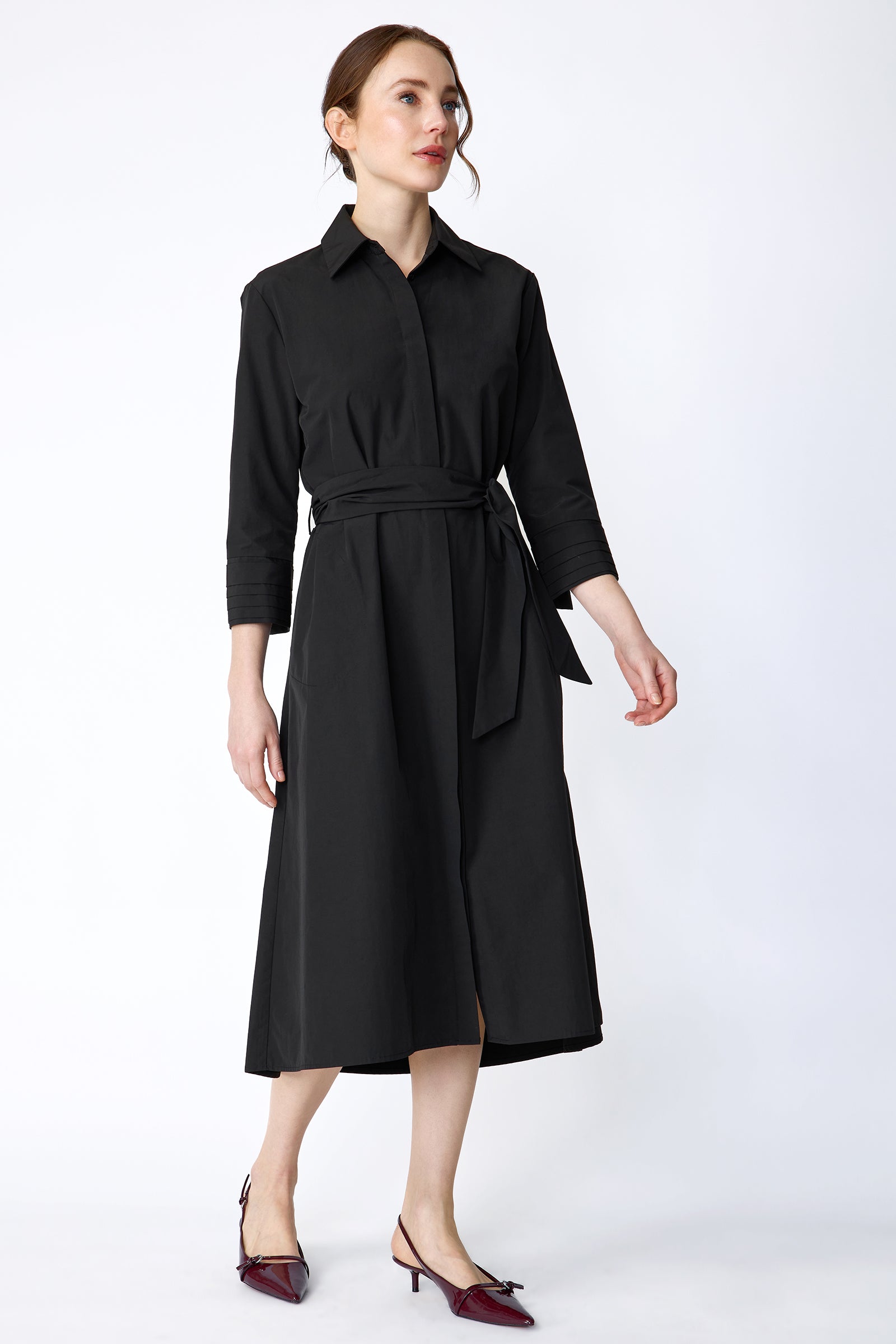 Woman wearing Kal Rieman Carolina Dress in Black Italian Broadcloth with burgundy shoes, against a white background, hand holding collar, arm on the side, looking sideways, walking, full front view