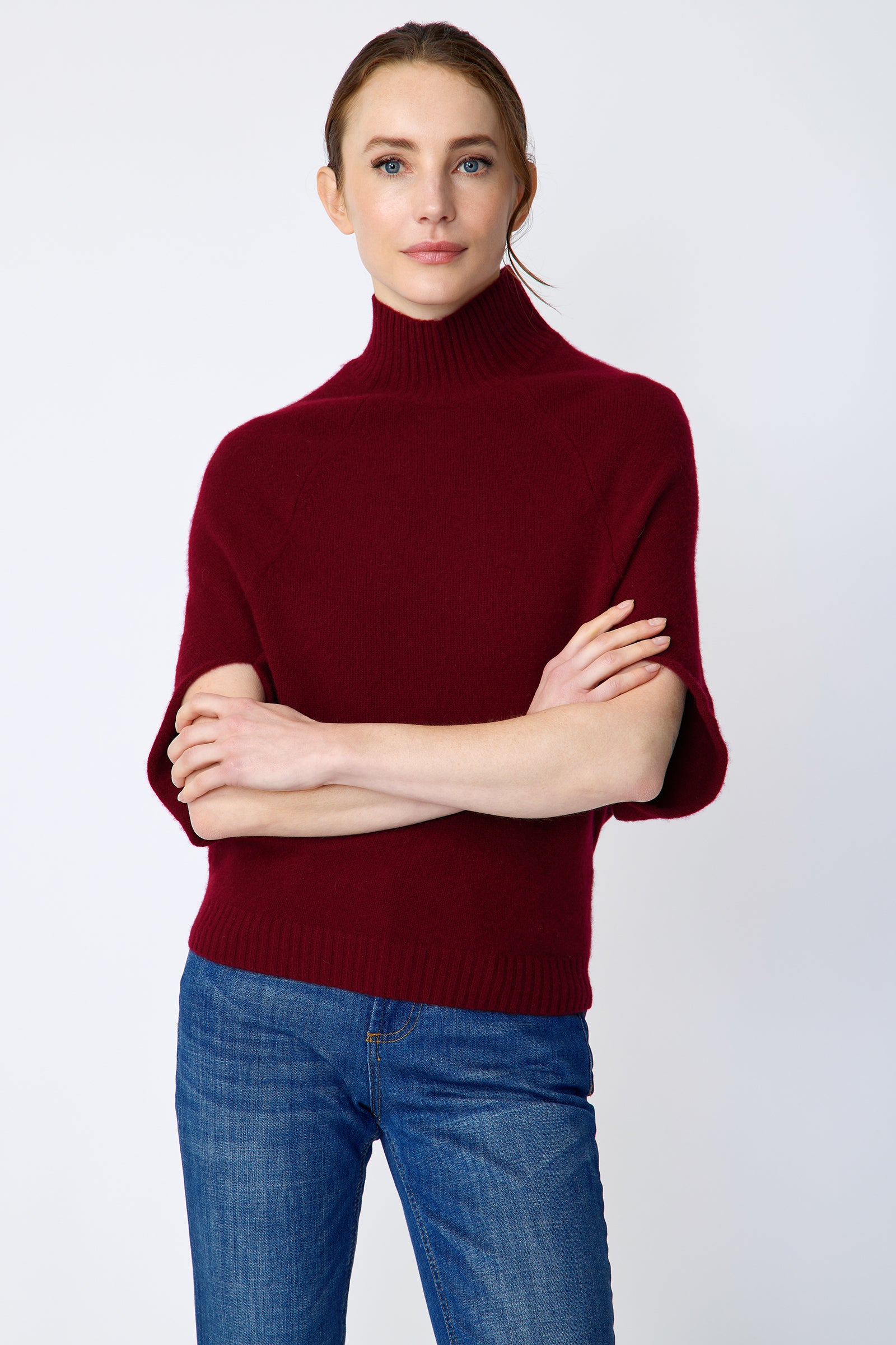 Woman wearing Kal Rieman bordeaux Cashmere Cape sweater and blue jeans on a white background arms crossed in front