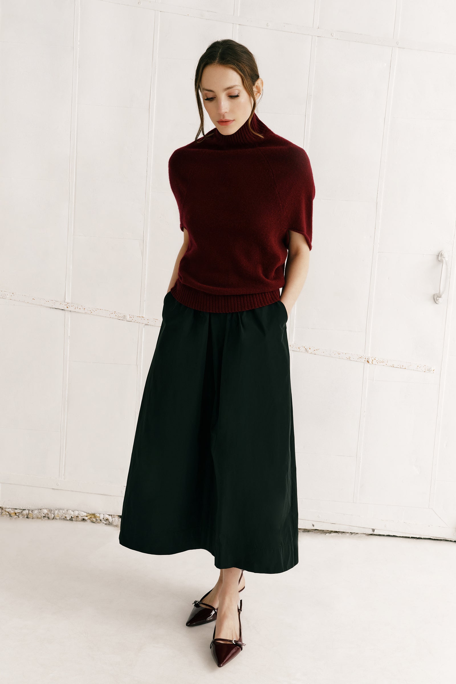 Woman wearing Kal Rieman Cashmere Cape Sweater in Bordeaux and Sadie skirt in black against a white wall arms in pockets look book image