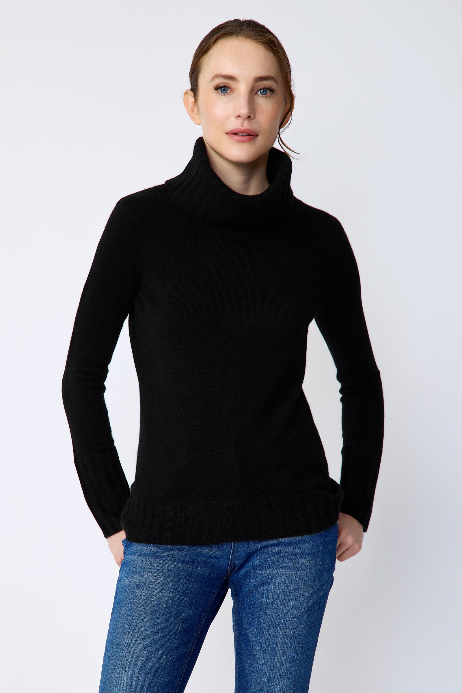Woman wearing a Kal Rieman Cashmere Cown Tneck in black sweater and blue jeans on a white background arms in pockets