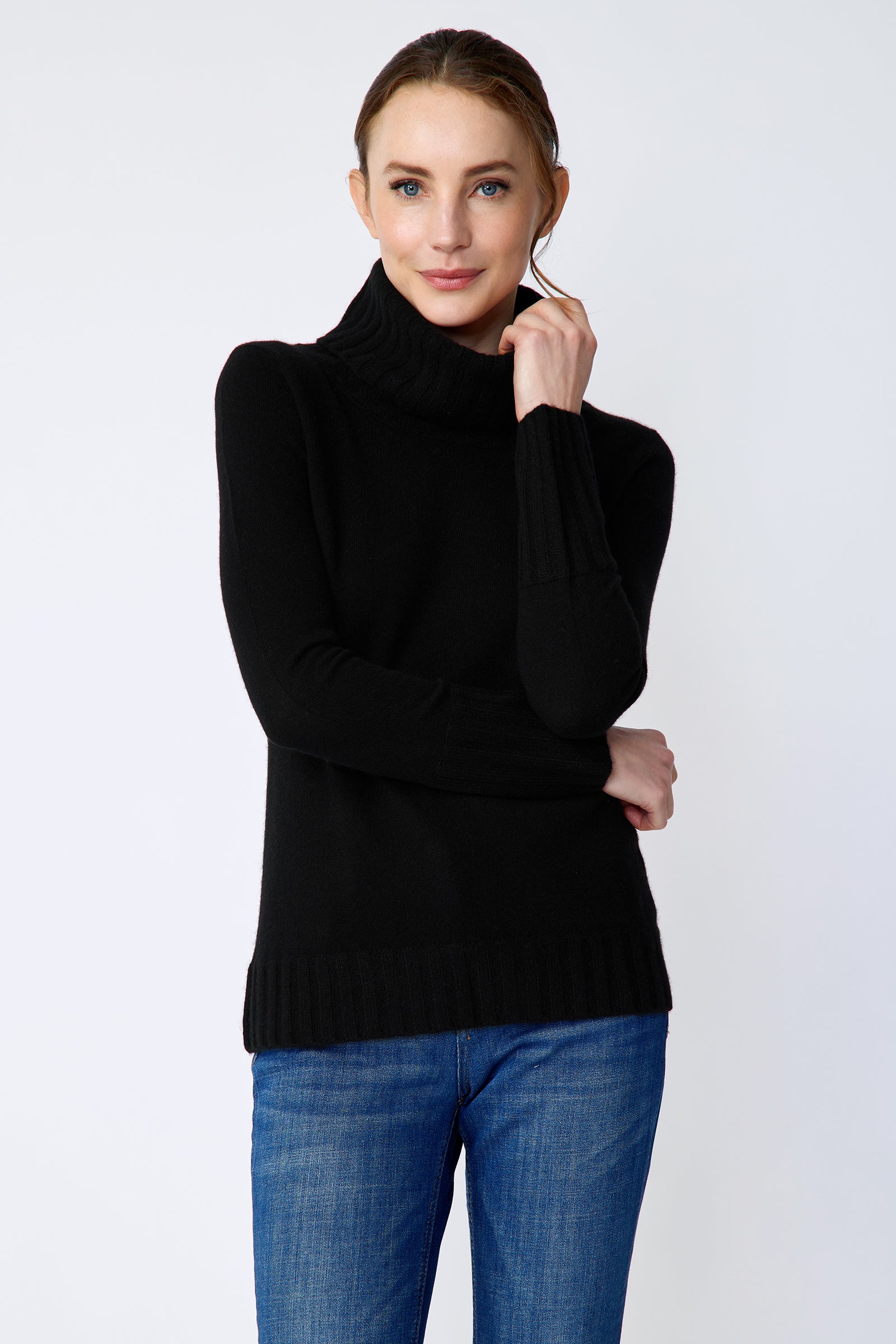 Woman wearing a Kal Rieman Cashmere Cown Tneck in black sweater and blue jeans on a white background arm crossed in front and holding collar