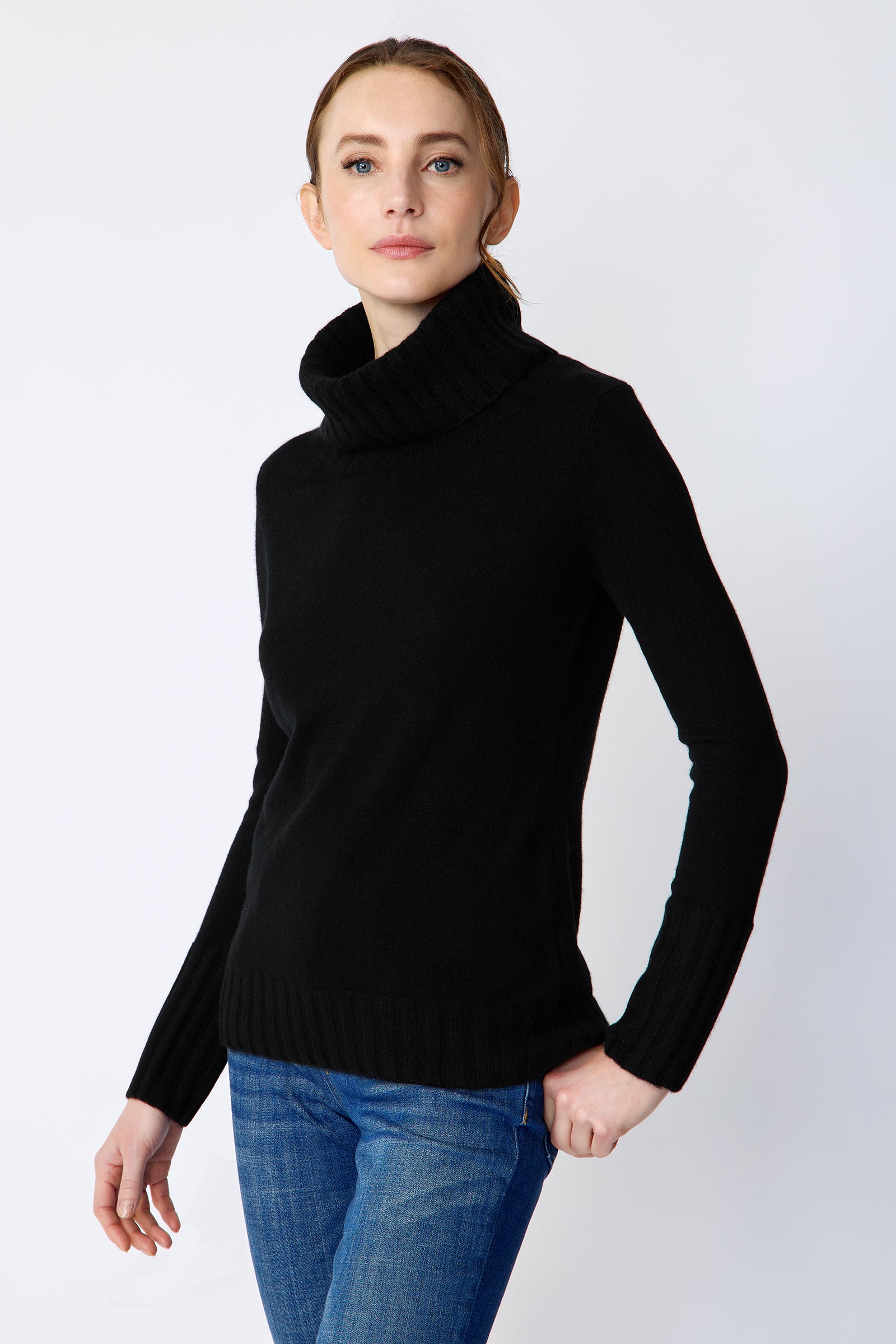 Woman wearing a Kal Rieman Cashmere Cown Tneck in black sweater and blue jeans on a white background arm in back pocket