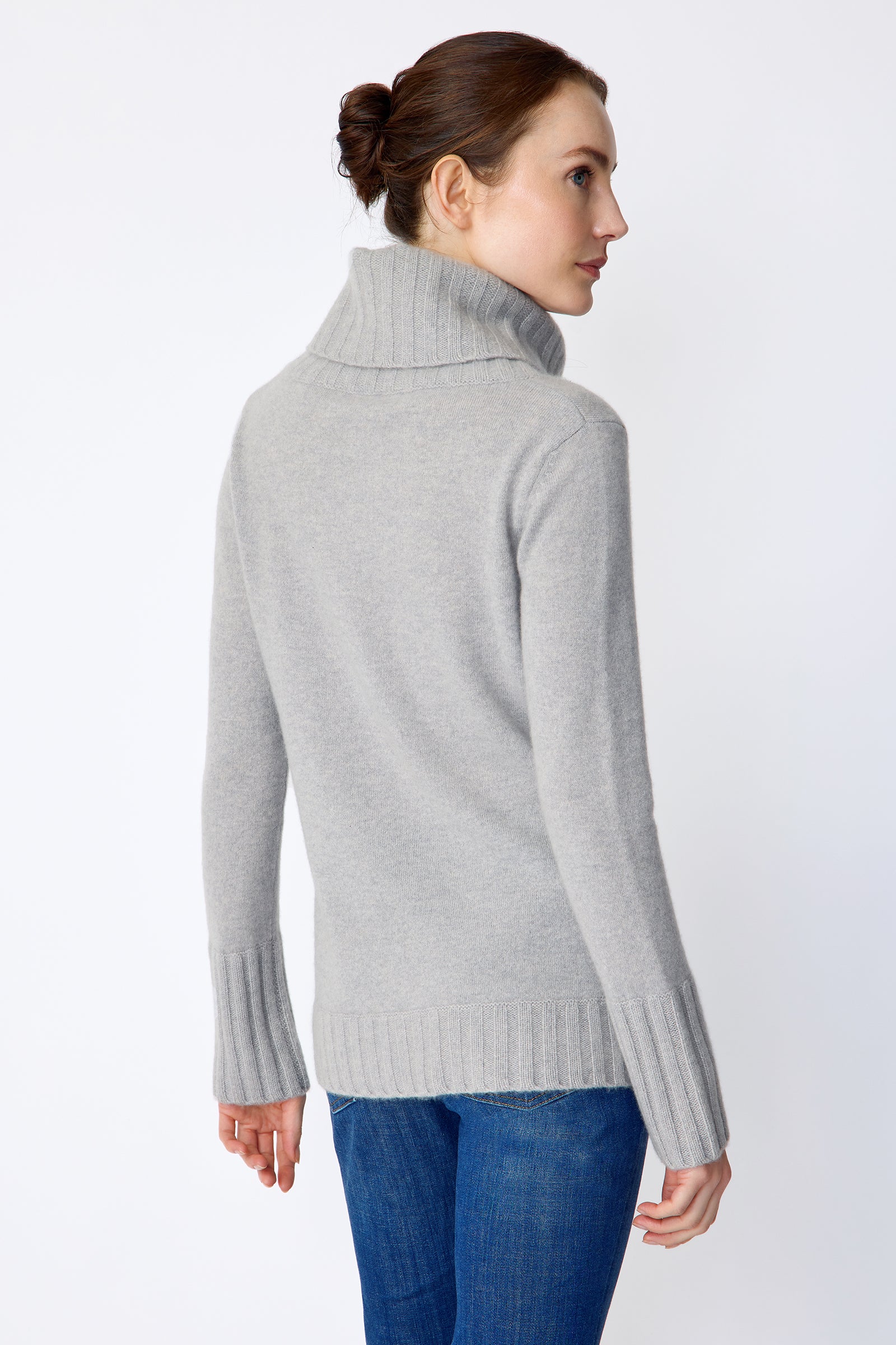 Woman wearing Kal Rieman Cashmere Cowl Tneck in Heather Grey and blue jeans on a white background arms on side back view