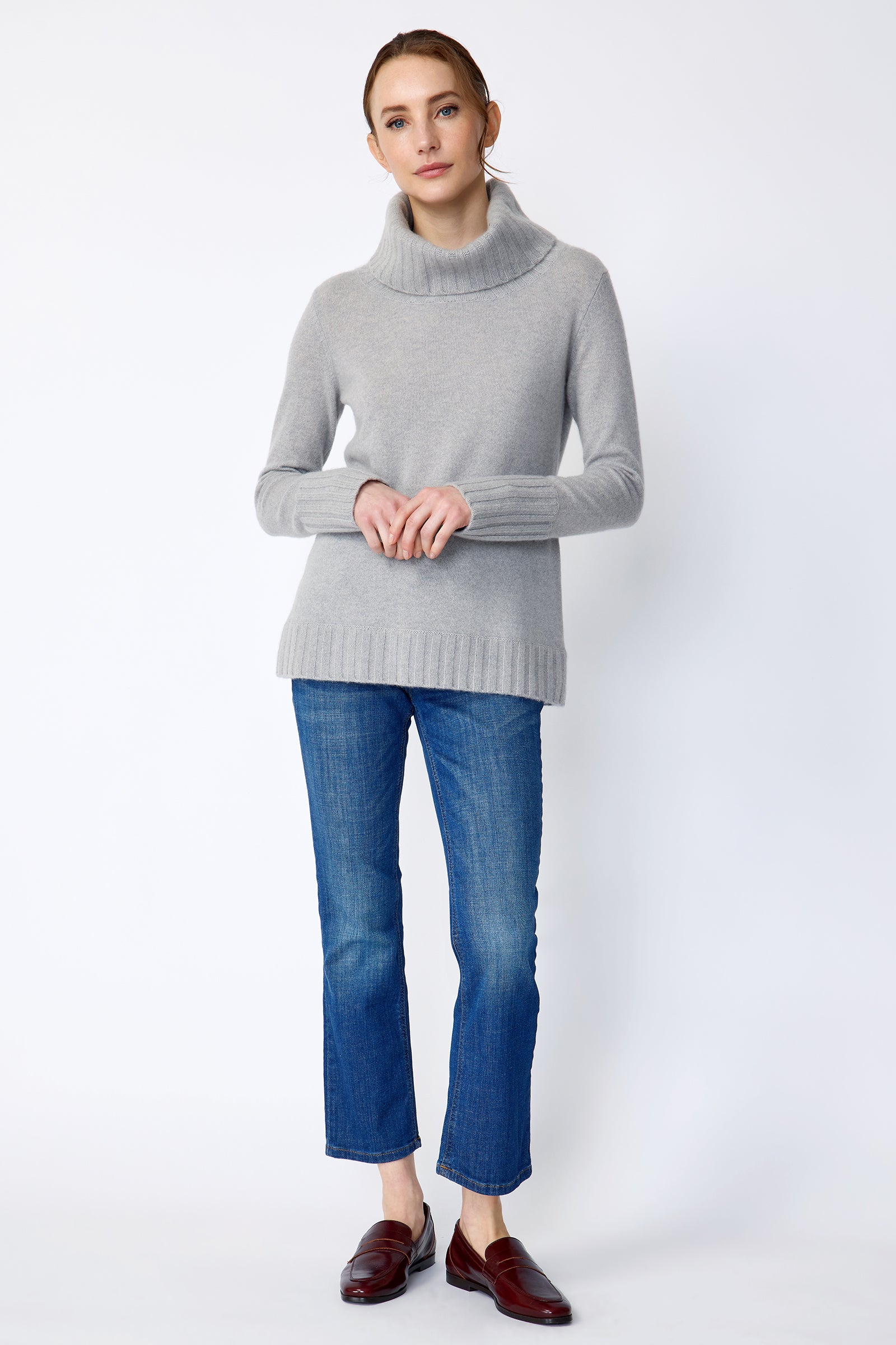 Woman wearing Kal Rieman Cashmere Cowl Tneck in Heather Grey and blue jeans on a white background holding hands in front