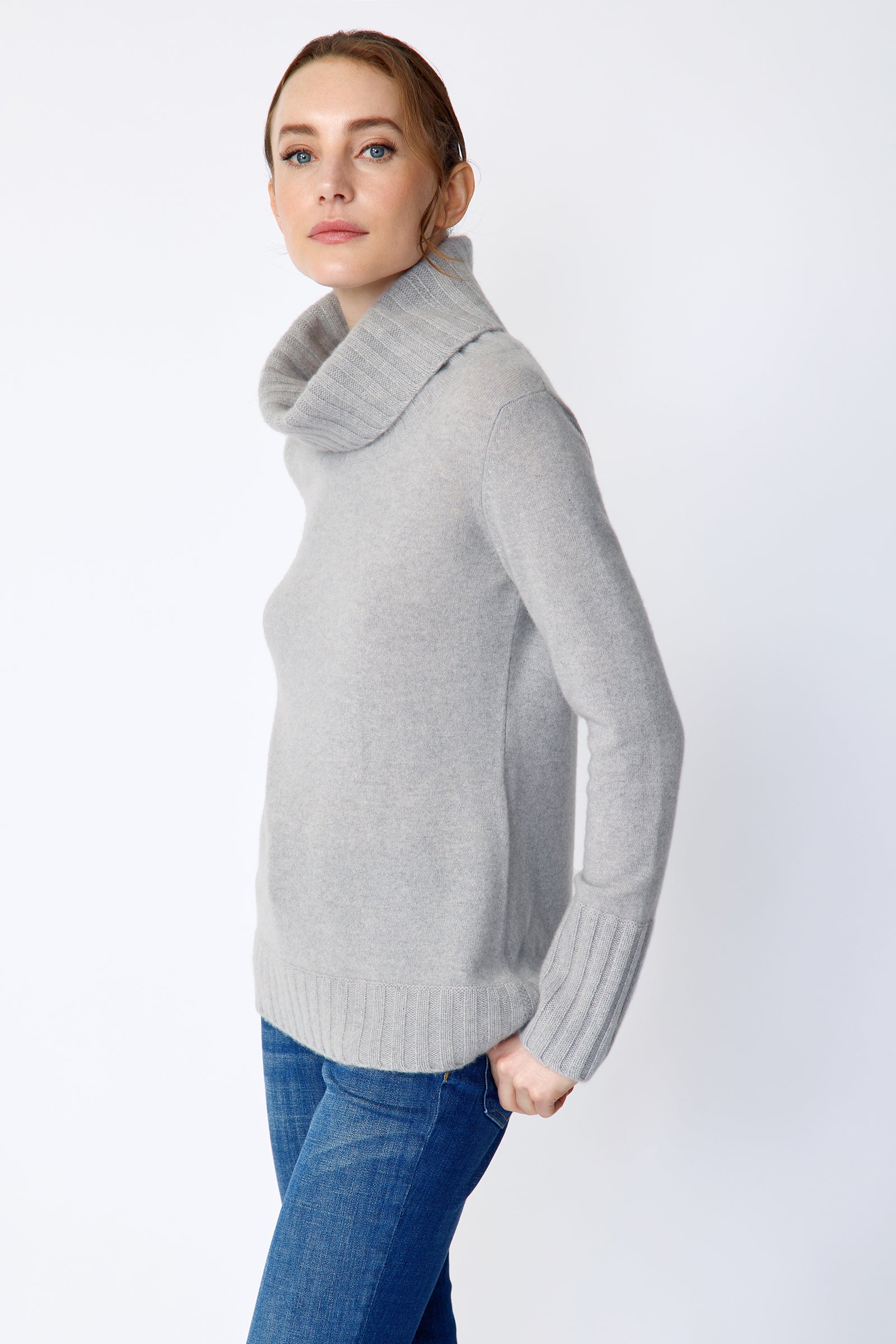 Woman wearing Kal Rieman Cashmere Cowl Tneck in Heather Grey and blue jeans on a white background arms in back pocket, side view