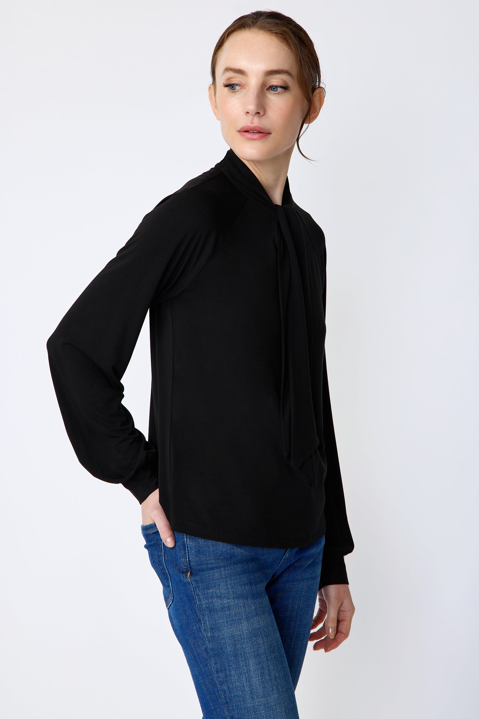 Woman wearing Kal Rieman Cecilia Scarf Top in Black and blue jeans on a white background, arm on the side, hand in pant  back pocket, side view