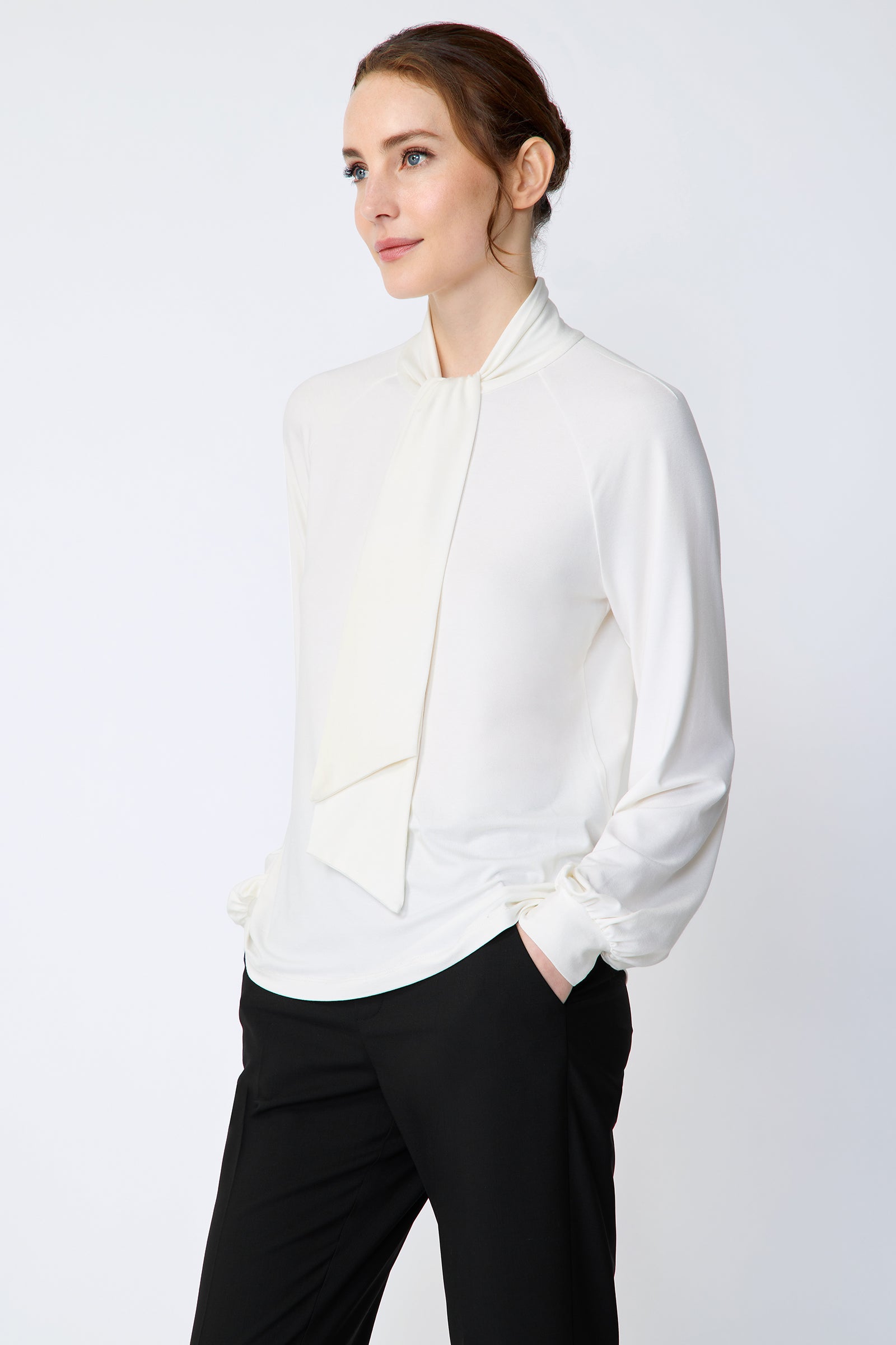 Woman wearing Kal Rieman Cecilia Scarf Top in Ivory and black pants on a white background, hands in pockets, side view
