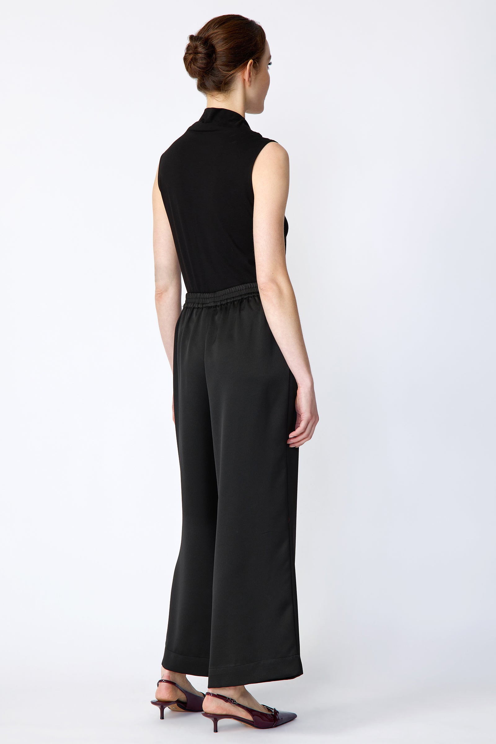 Woman wearing Kal Rieman Celeste pant in Black poly satin with black sleeveless tank on a white background, hands in pockets, full front view