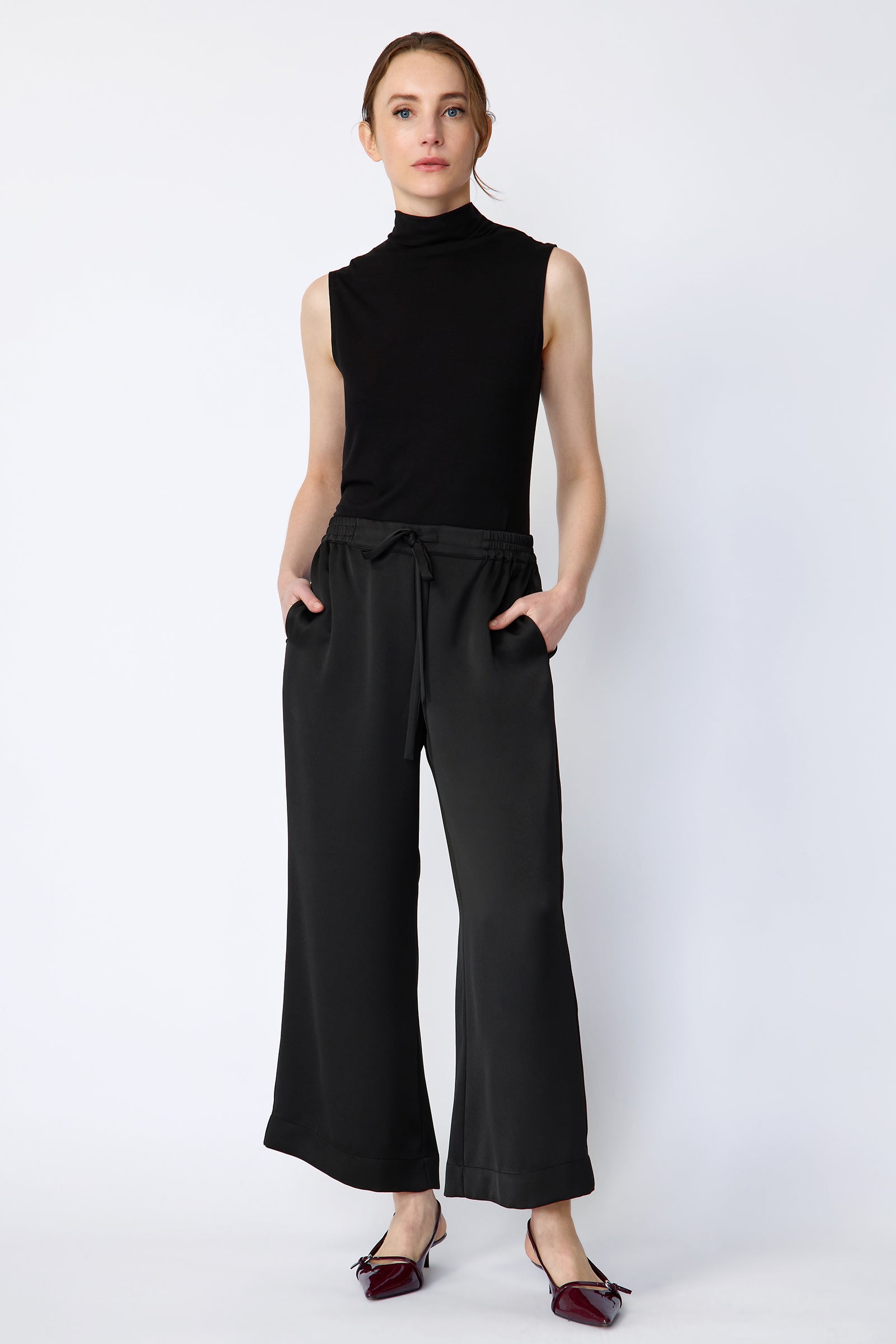 Woman wearing Kal Rieman Celeste pant in Black poly satin with black sleeveless tank on a white background, hands in pockets, full front view