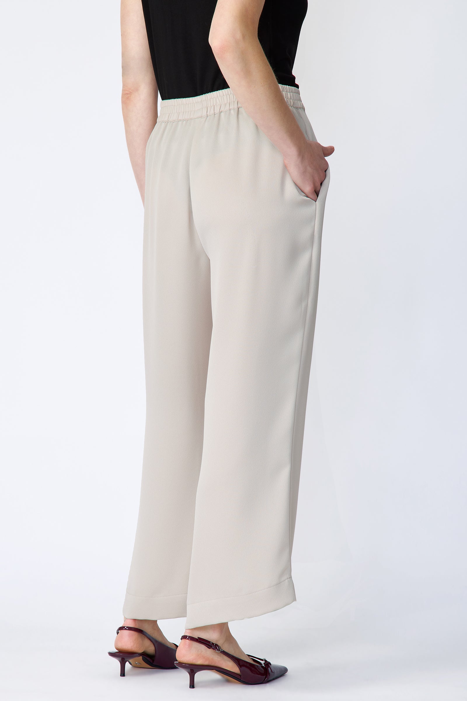 Woman wearing Kal Rieman Celeste Pant in Mink Poly Satin with a black sleeveless top on a white background, hands in pant pockets, full front view