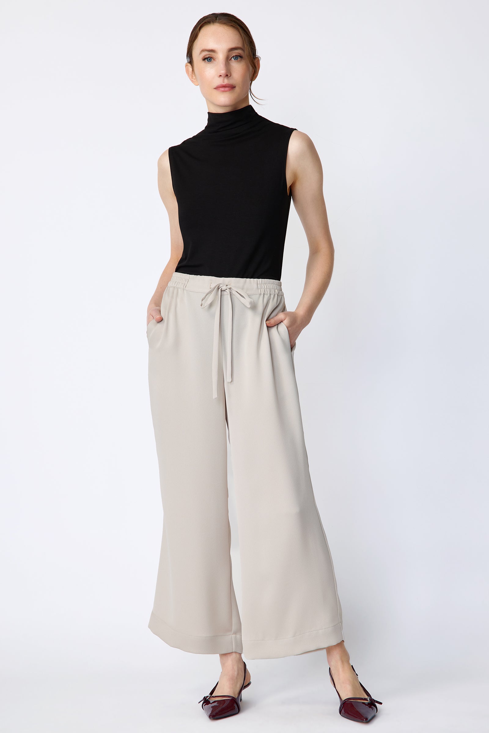 Woman wearing Kal Rieman Celeste Pant in Mink Poly Satin with a black sleeveless top on a white background, hands in pant pockets, full front view