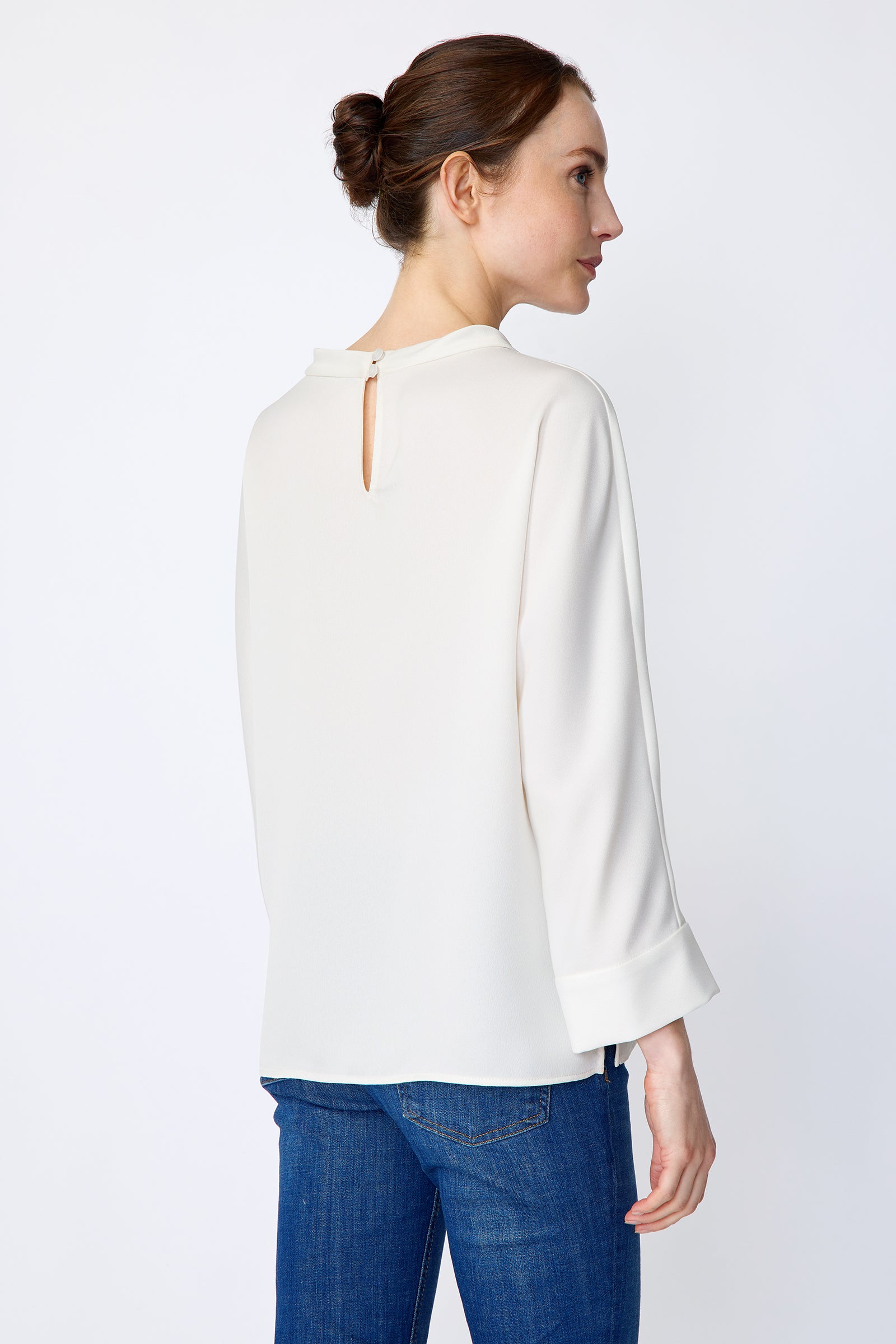 Woman wearing Kal Rieman Elena Drape Top in Ivory and blue jeans on a white background, arms on the side, back view