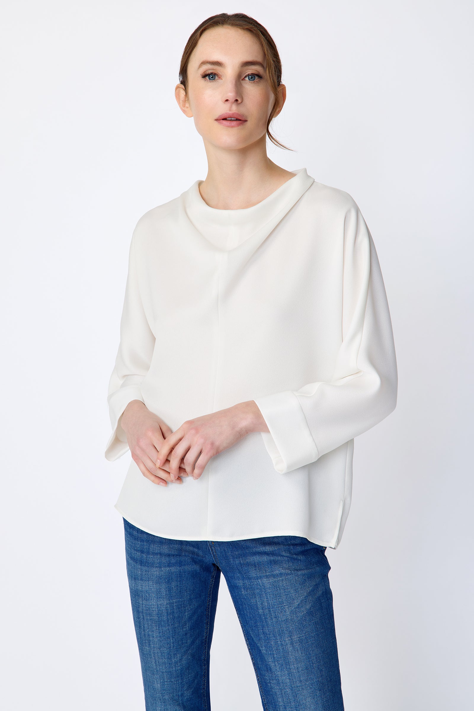 Woman wearing Kal Rieman Elena Drape Top in Ivory and blue jeans on a white background, holdings hands in front, close view
