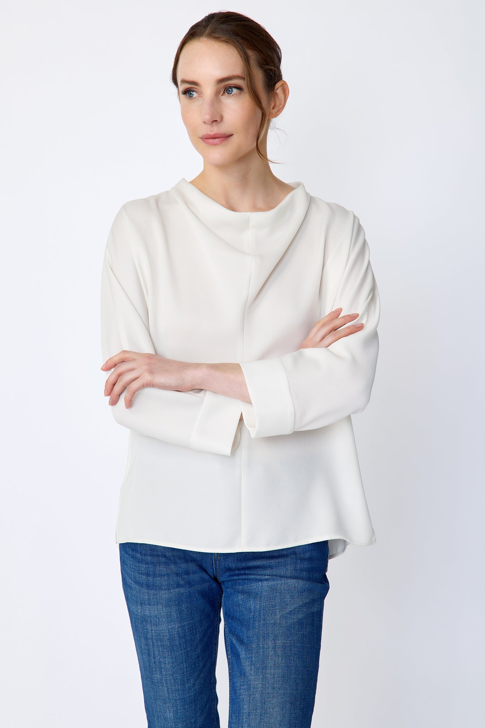 Woman wearing Kal Rieman Elena Drape Top in Ivory and blue jeans on a white background, arms crossed in front, close view