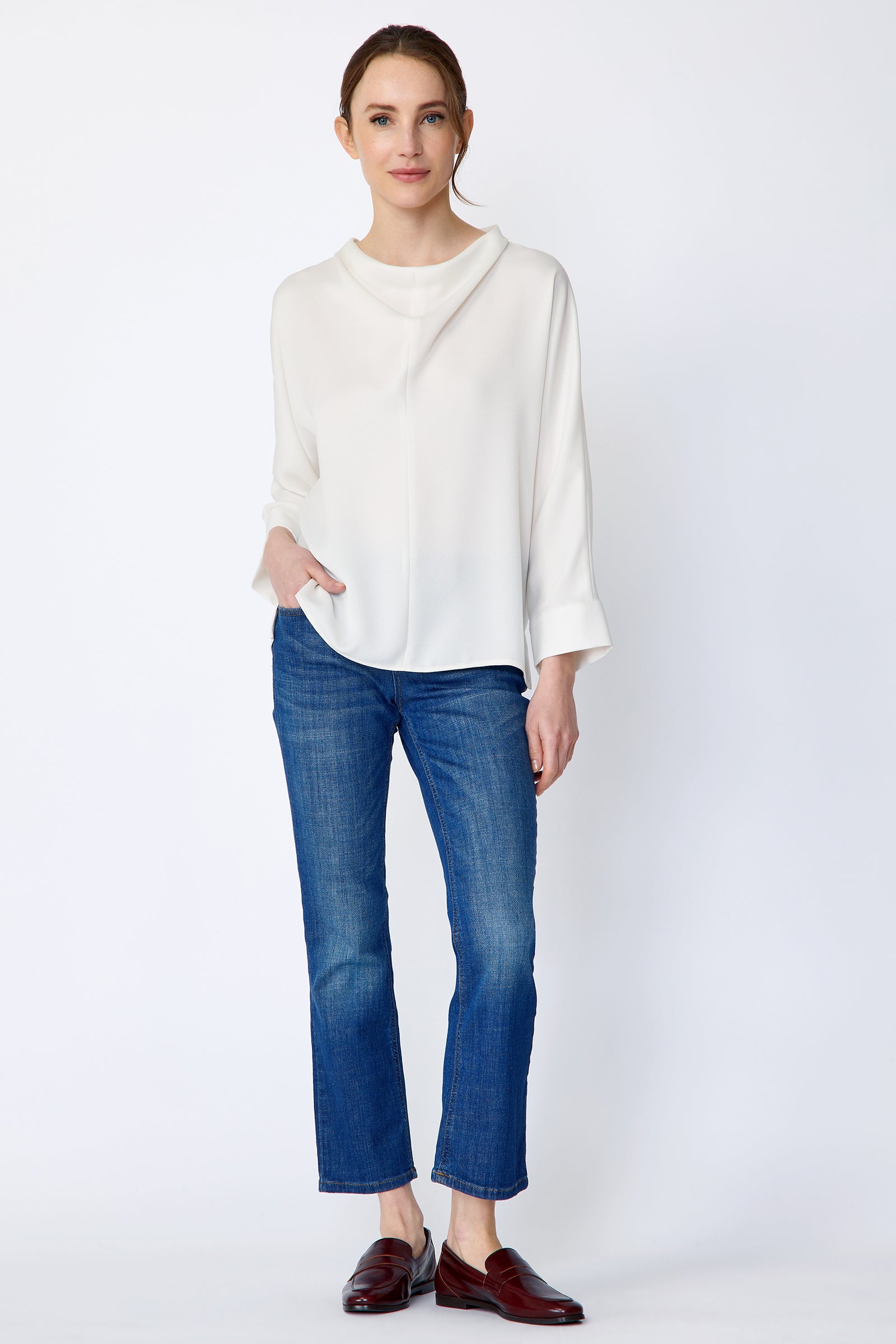 Woman wearing Kal Rieman Elena Drape Top in Ivory and blue jeans on a white background, hand in pant pocket, arm on the side