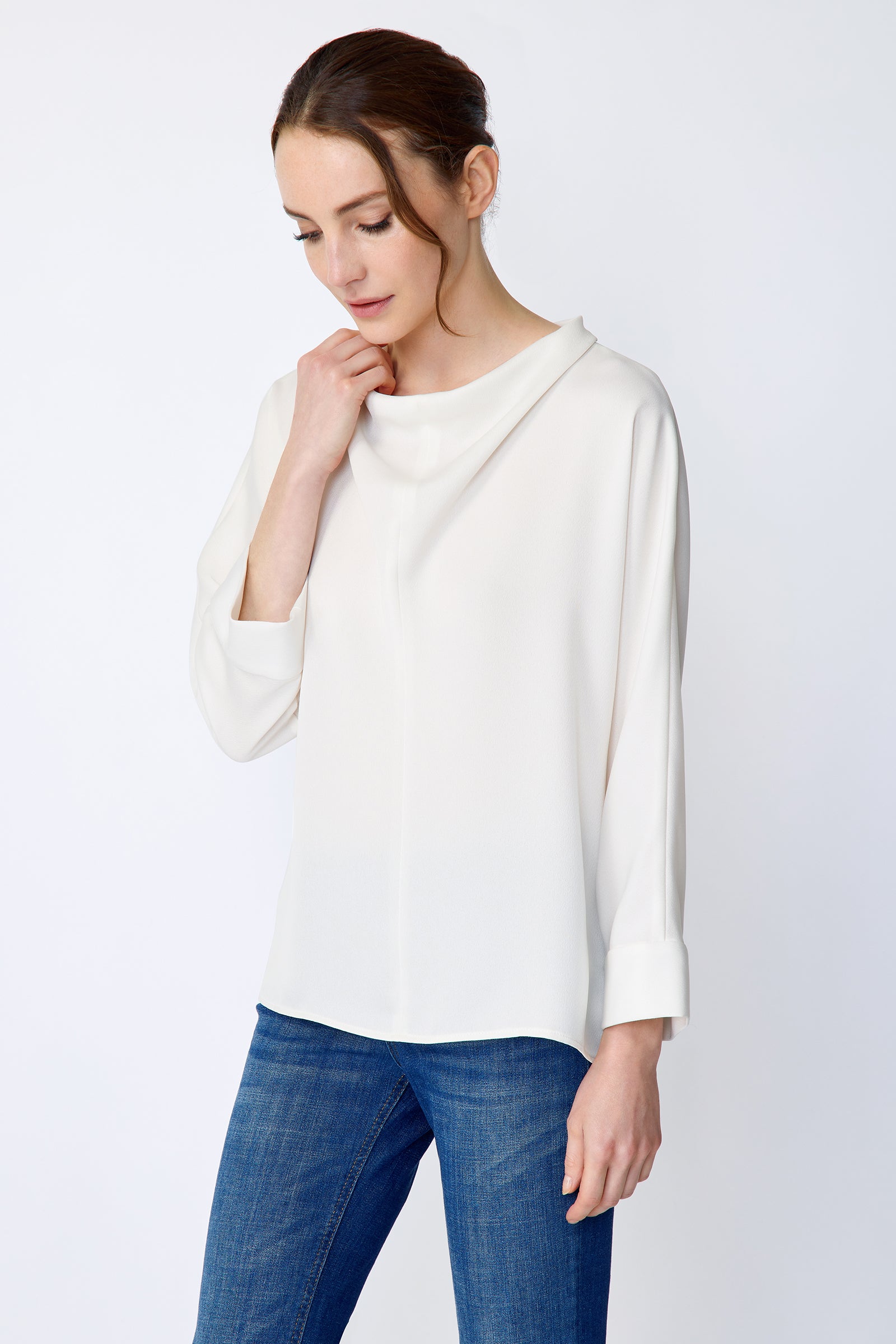 Woman wearing Kal Rieman Elena Drape Top in Ivory and blue jeans on a white background, holding collar, arm on the side, looking down