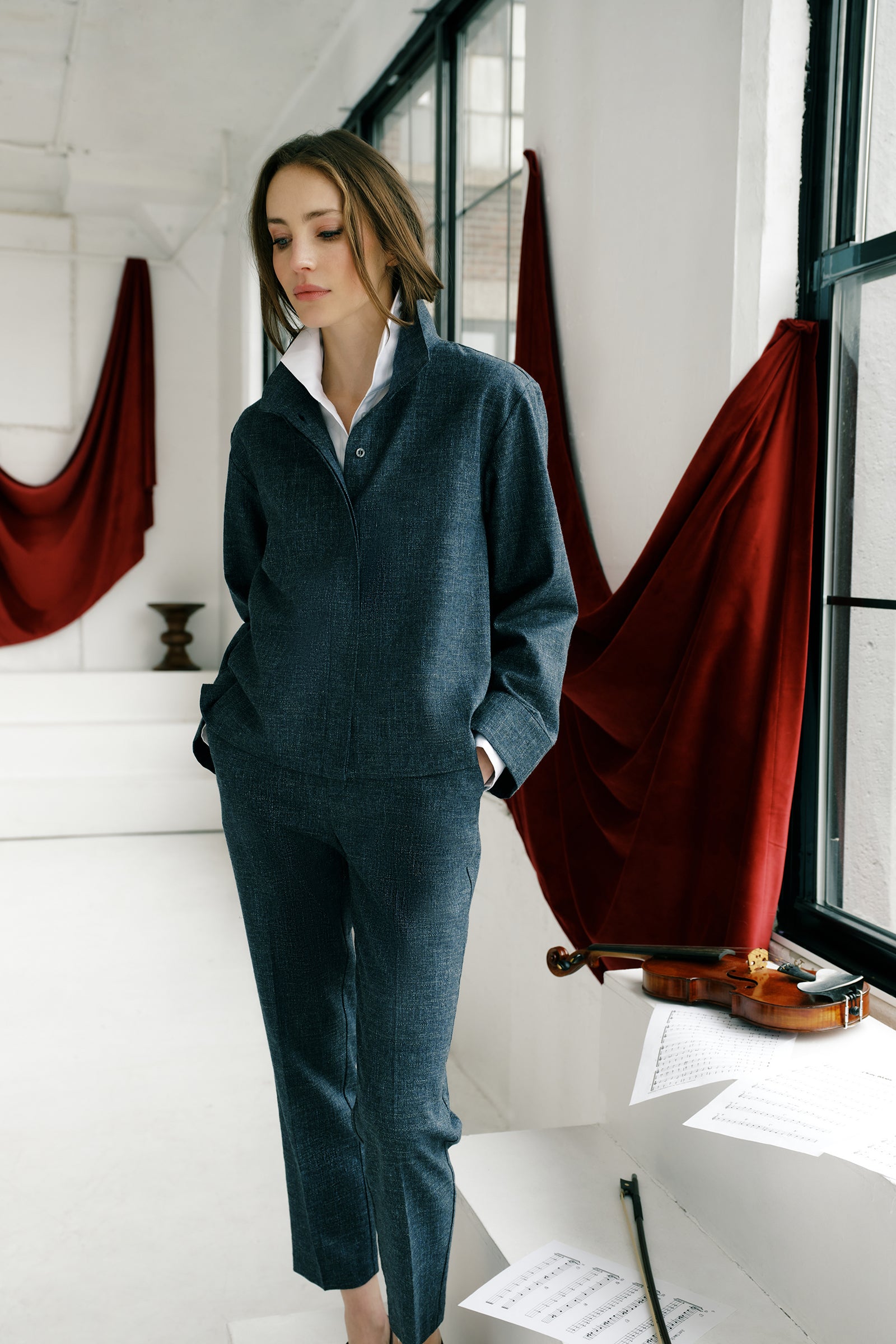 Woman wearing a Kal Rieman Morgan Shirt Jacket in Denim with the Kal Rieman Martine Trouser in Denim in a room with red curtain drapes, steps with a violin and sheet music, hands in pant pockets look book image