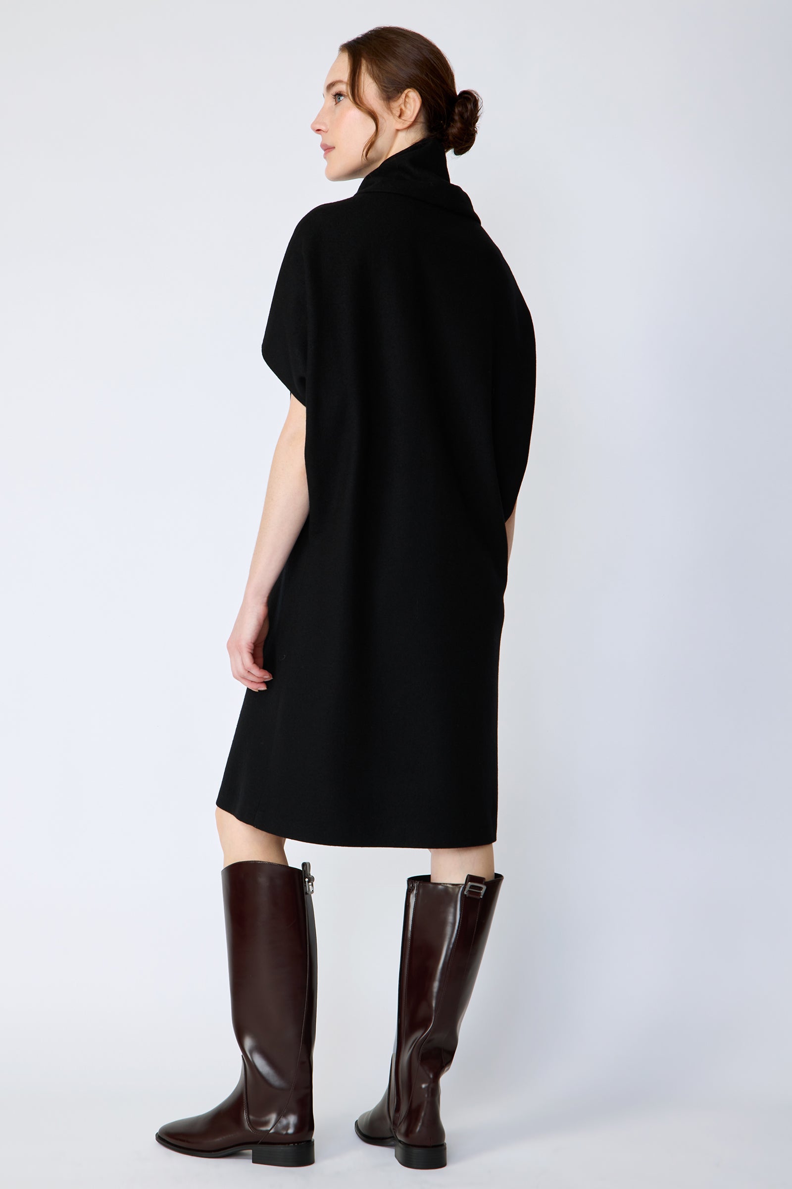 Woman wearing Kal Rieman Nina Cape Dress in Black with brown knee high boots on a white background, arms on the side, back view
