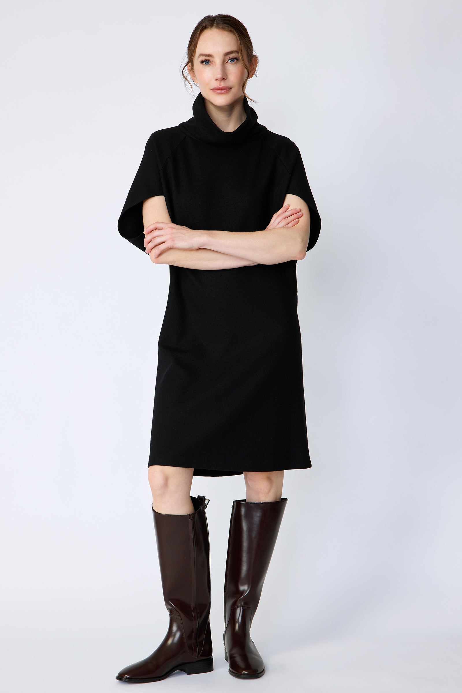 Woman wearing Kal Rieman Nina Cape Dress in Black with brown knee high boots on a white background, arms crossed in the front, full front view