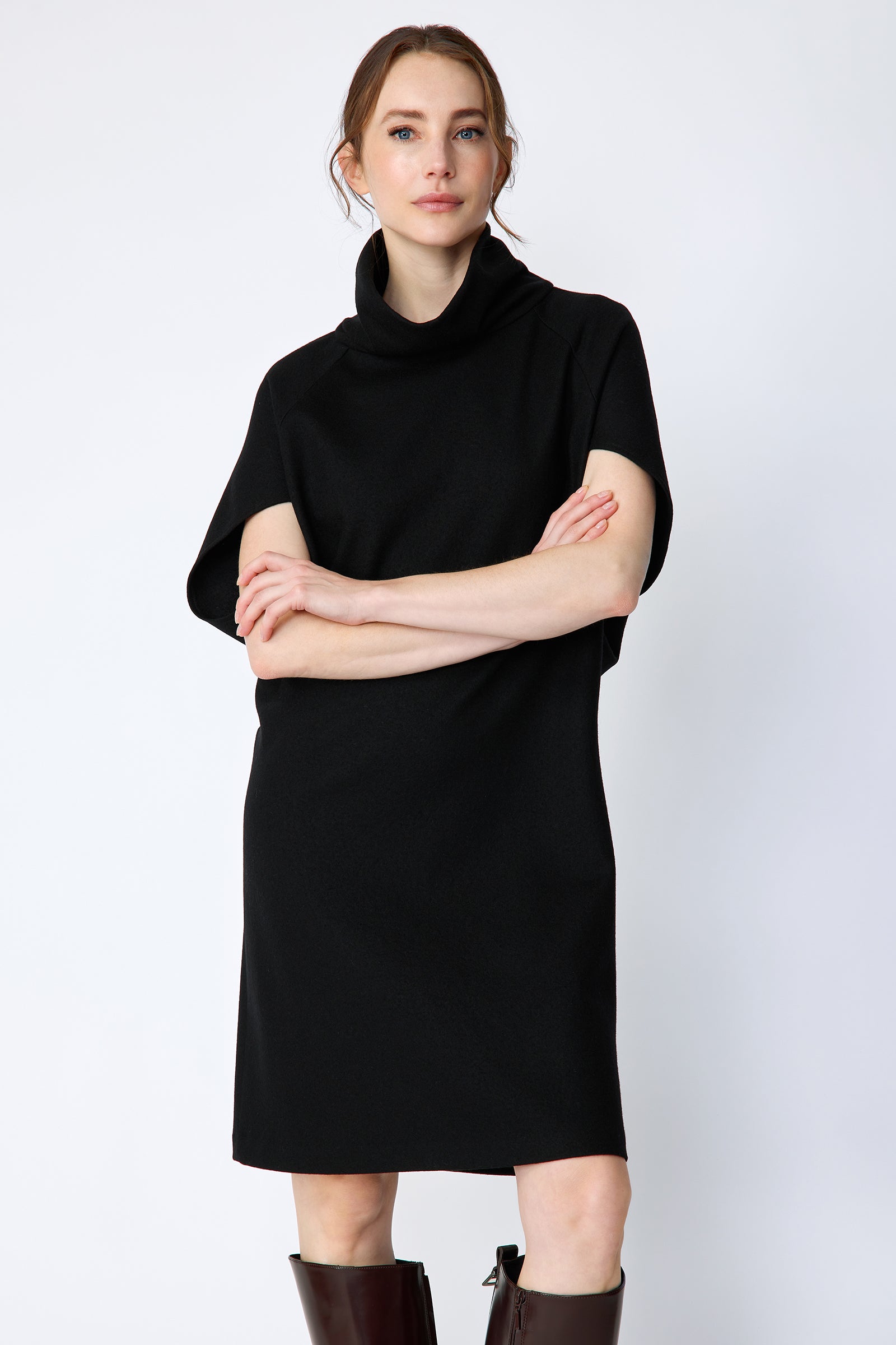 Woman wearing Kal Rieman Nina Cape Dress in Black with brown knee high boots on a white background, arms crossed in the front, closeview