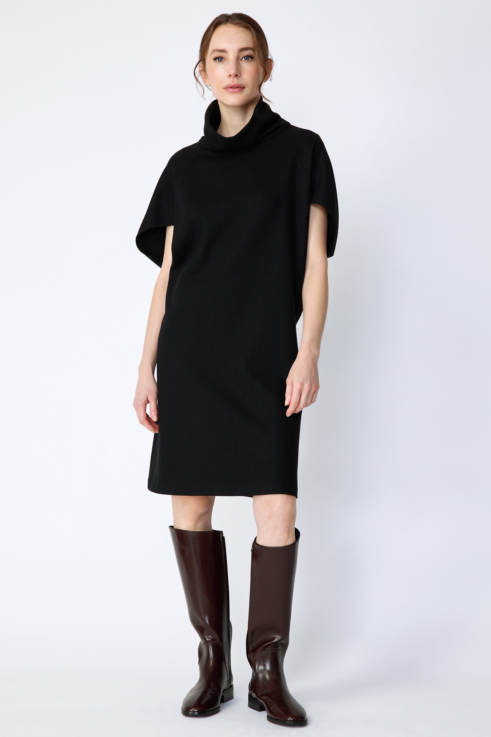 Woman wearing Kal Rieman Nina Cape Dress in Black with brown knee high boots on a white background, arms on the side, full front view
