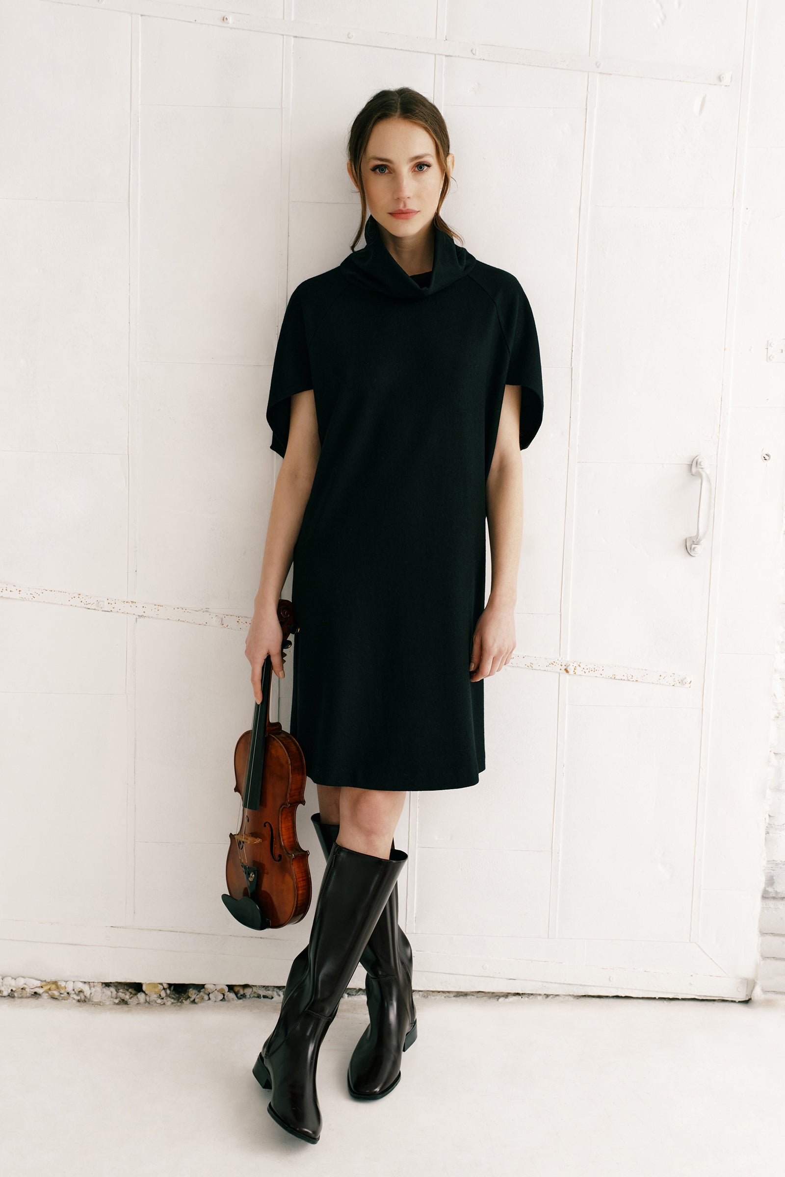 Woman wearing Kal Rieman Nina Cape Dress in Black with brown knee high boots on a white background, arms crossed in the front, full front view