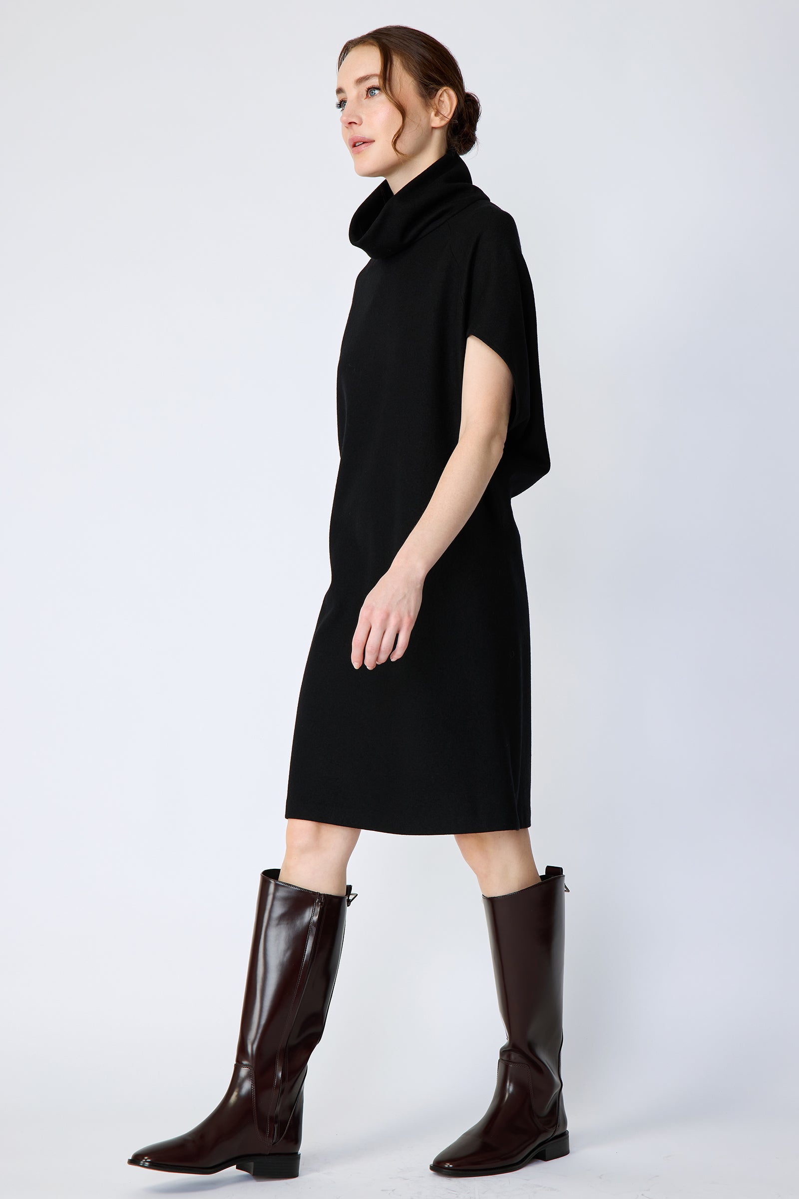 Woman wearing Kal Rieman Nina Cape Dress in Black with brown knee high boots on a white background, arms on the side, walking, side view