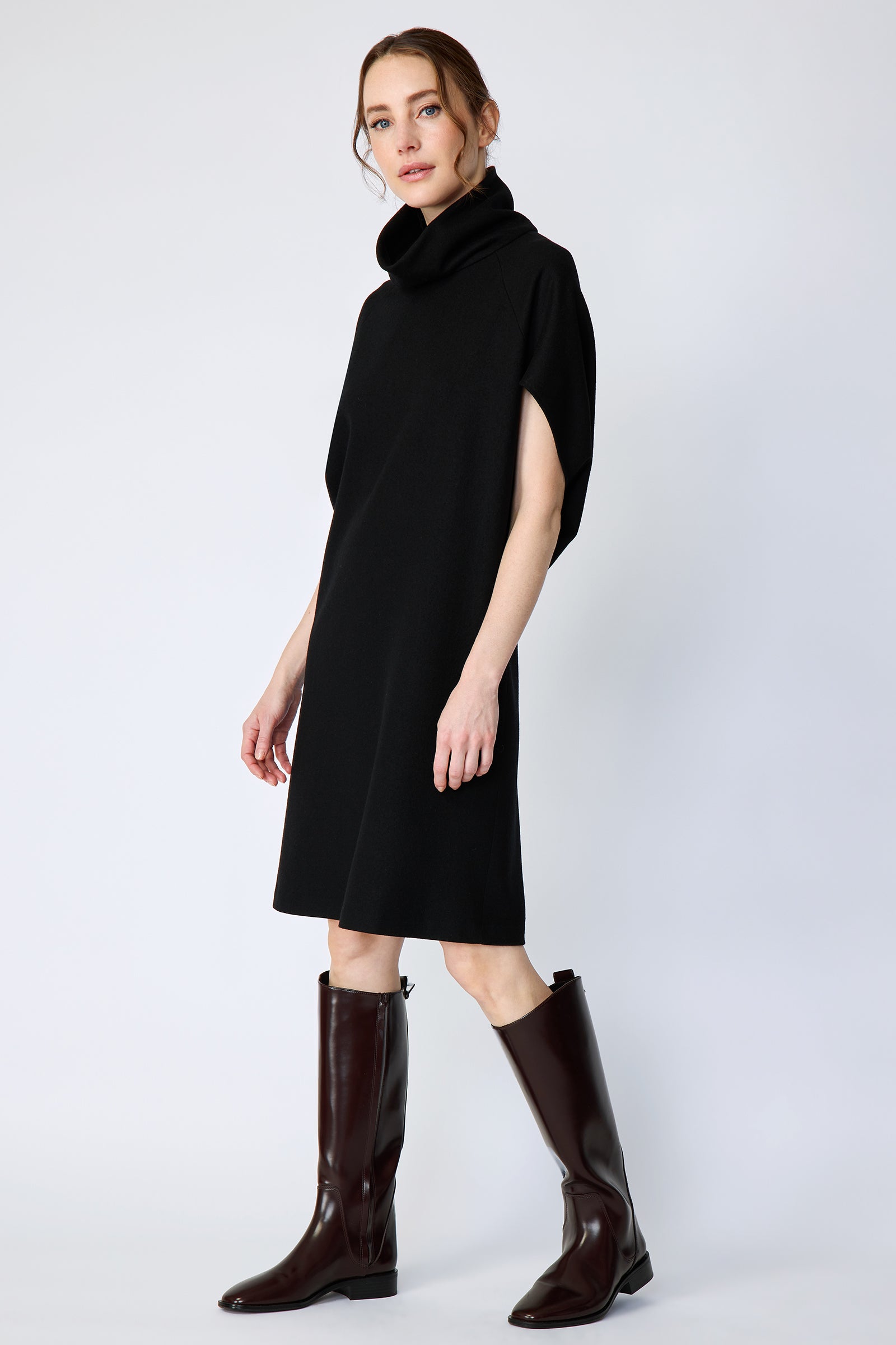 Woman wearing Kal Rieman Nina Cape Dress in Black with brown knee high boots on a white background, arms on the side, walking, side view