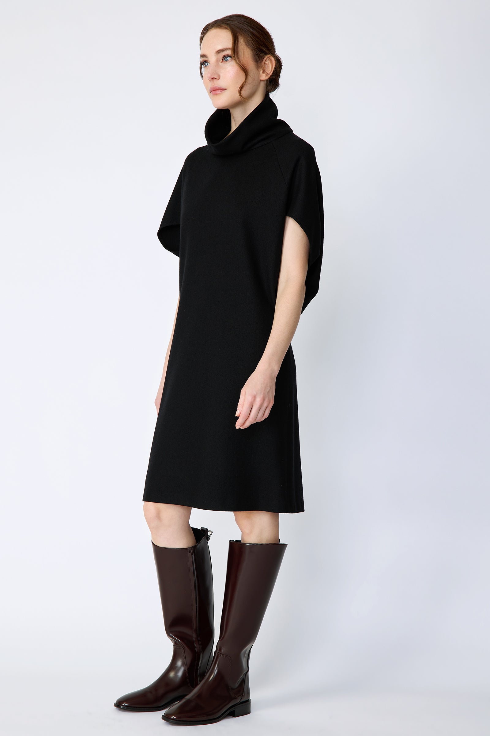 Woman wearing Kal Rieman Nina Cape Dress in Black with brown knee high boots on a white background, arms on the side, front view