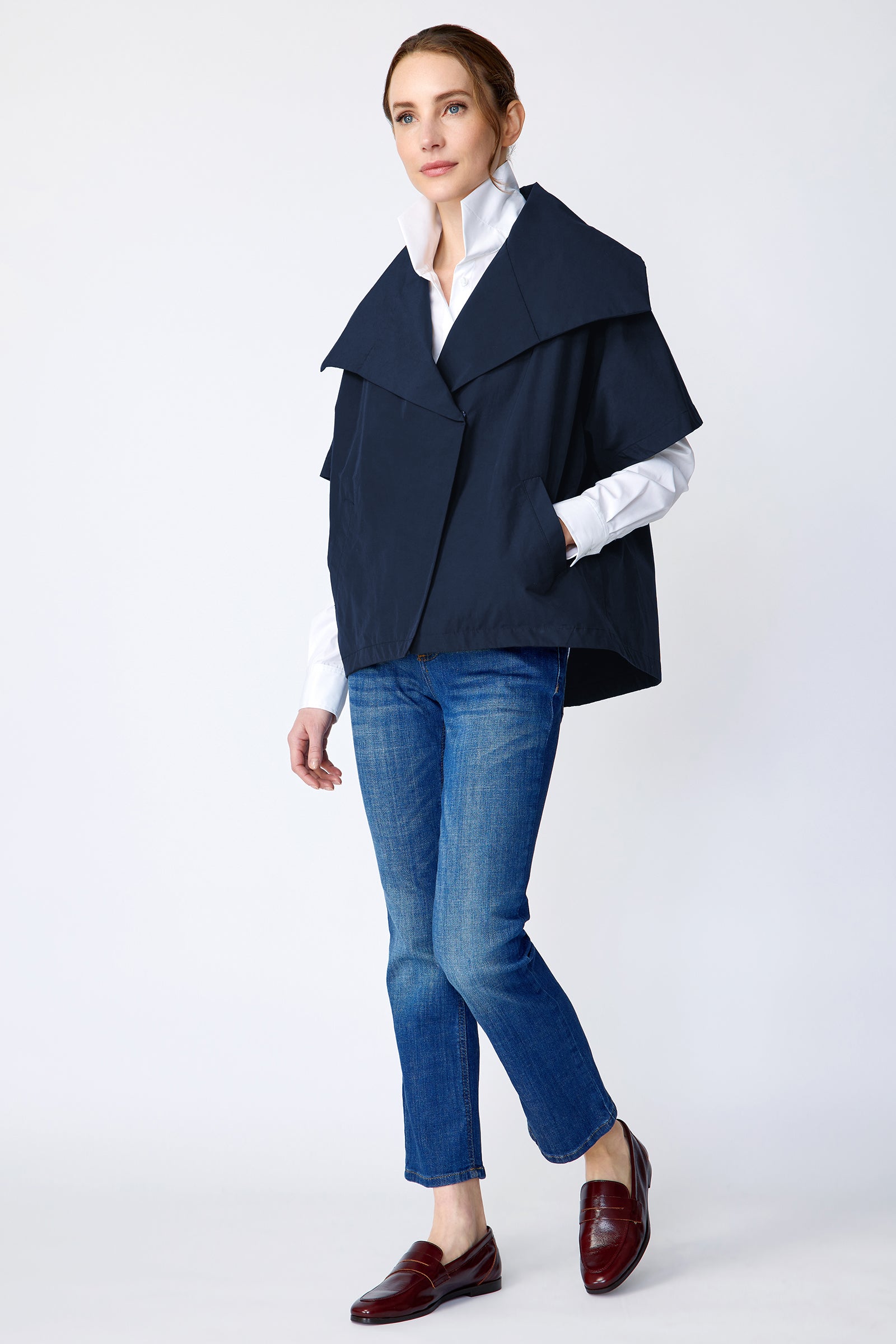 Woman wearing Kal Rieman navy blue Paris jacket, white shirt, and blue jeans on a white background side view walking with hand in pocket