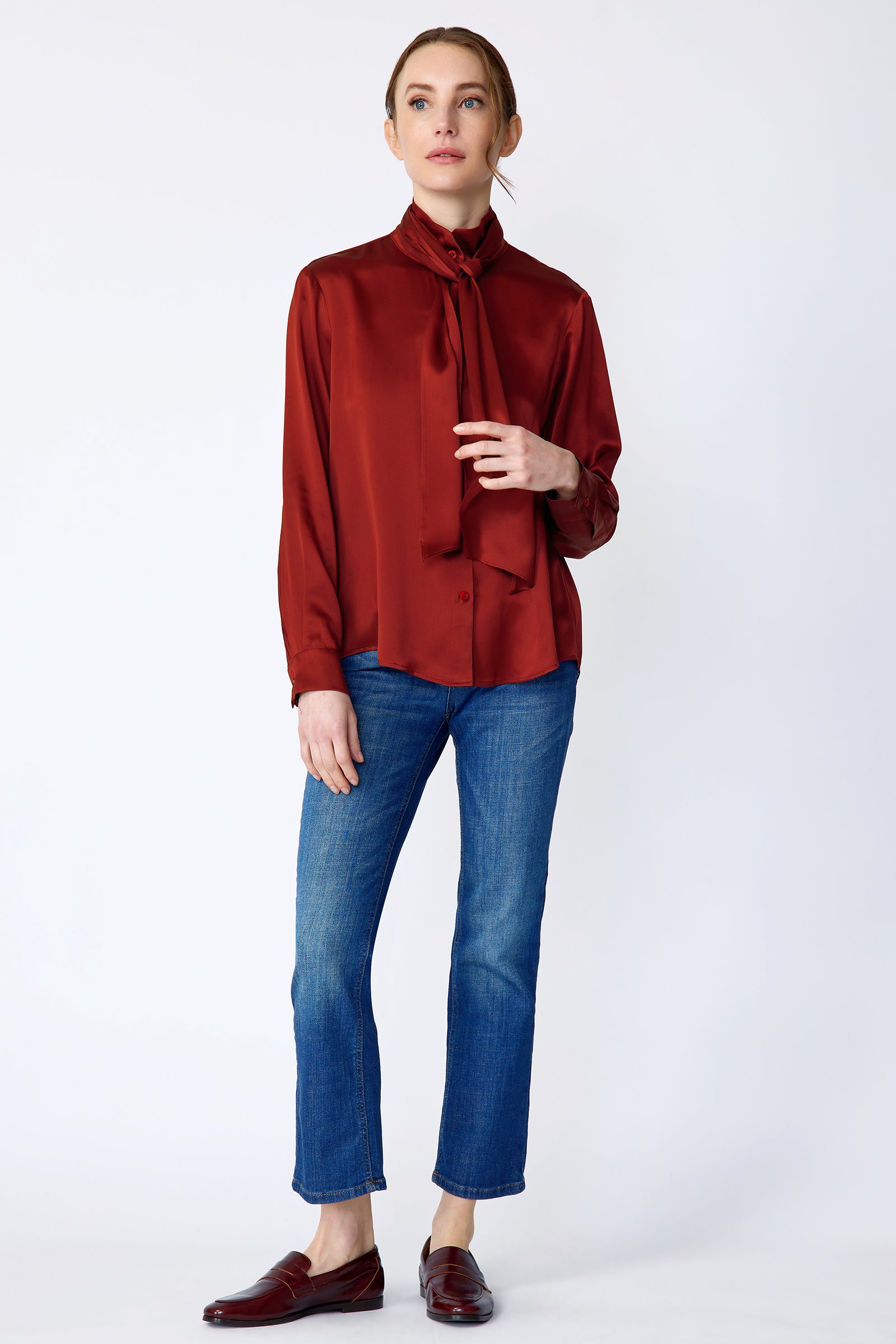 Woman wearing Kal Rieman Silk Regency Blouse in Rouge and blue jeans on a white background, holding scarf, arm on the side, full front view