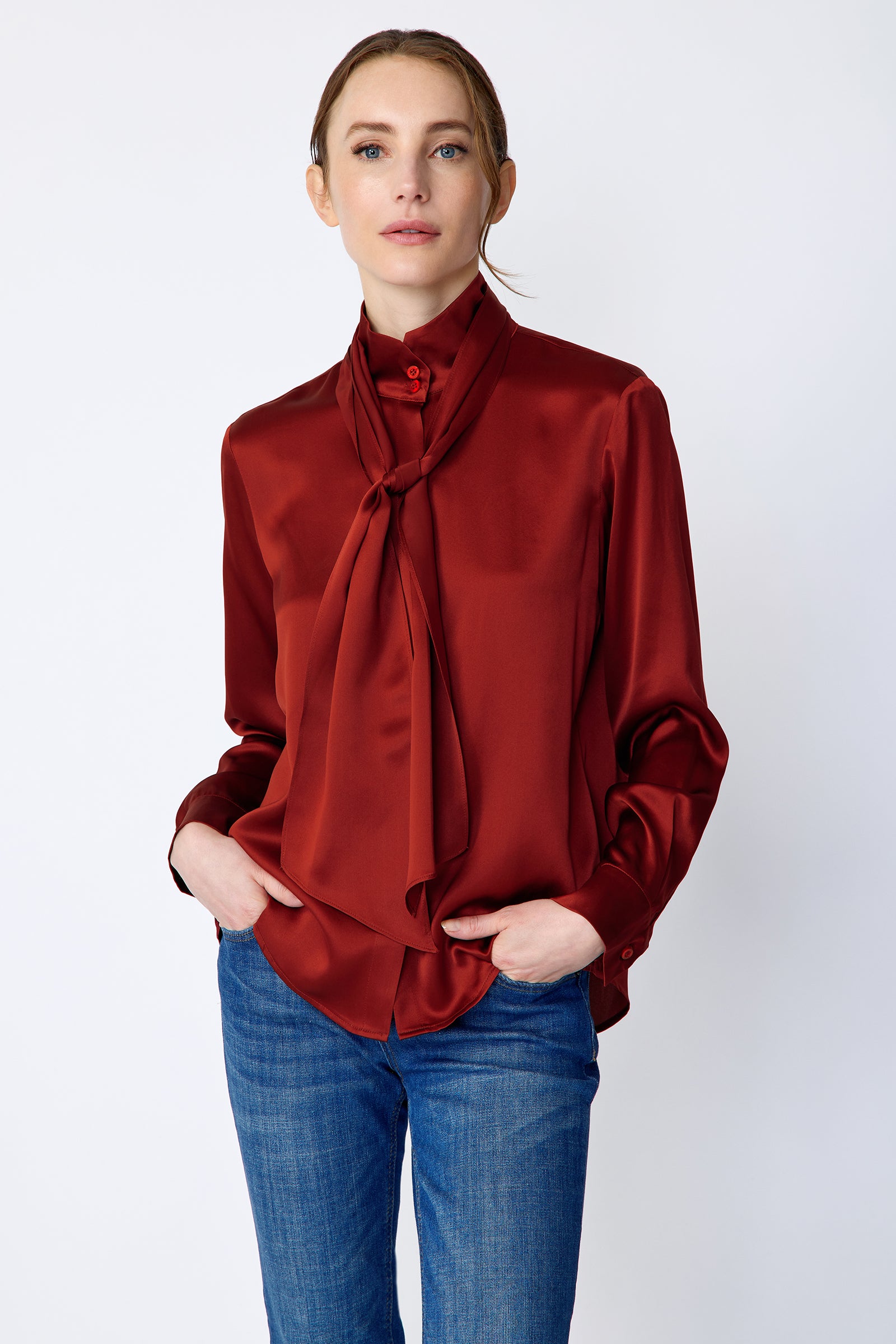 Woman wearing Kal Rieman Silk Regency Blouse in Rouge and blue jeans on a white background, hands in pant front pockets, close view