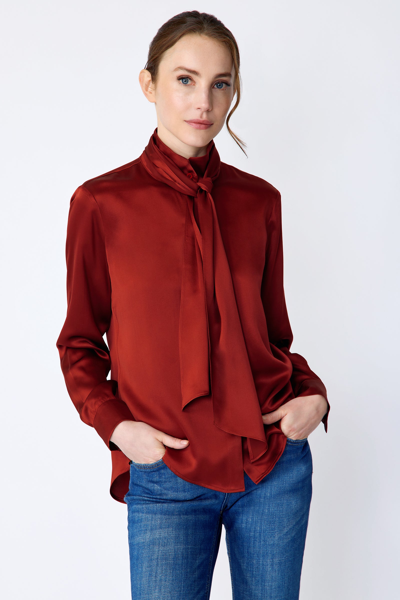Woman wearing Kal Rieman Silk Regency Blouse in Rouge and blue jeans on a white background, hands in pant front pockets, close view