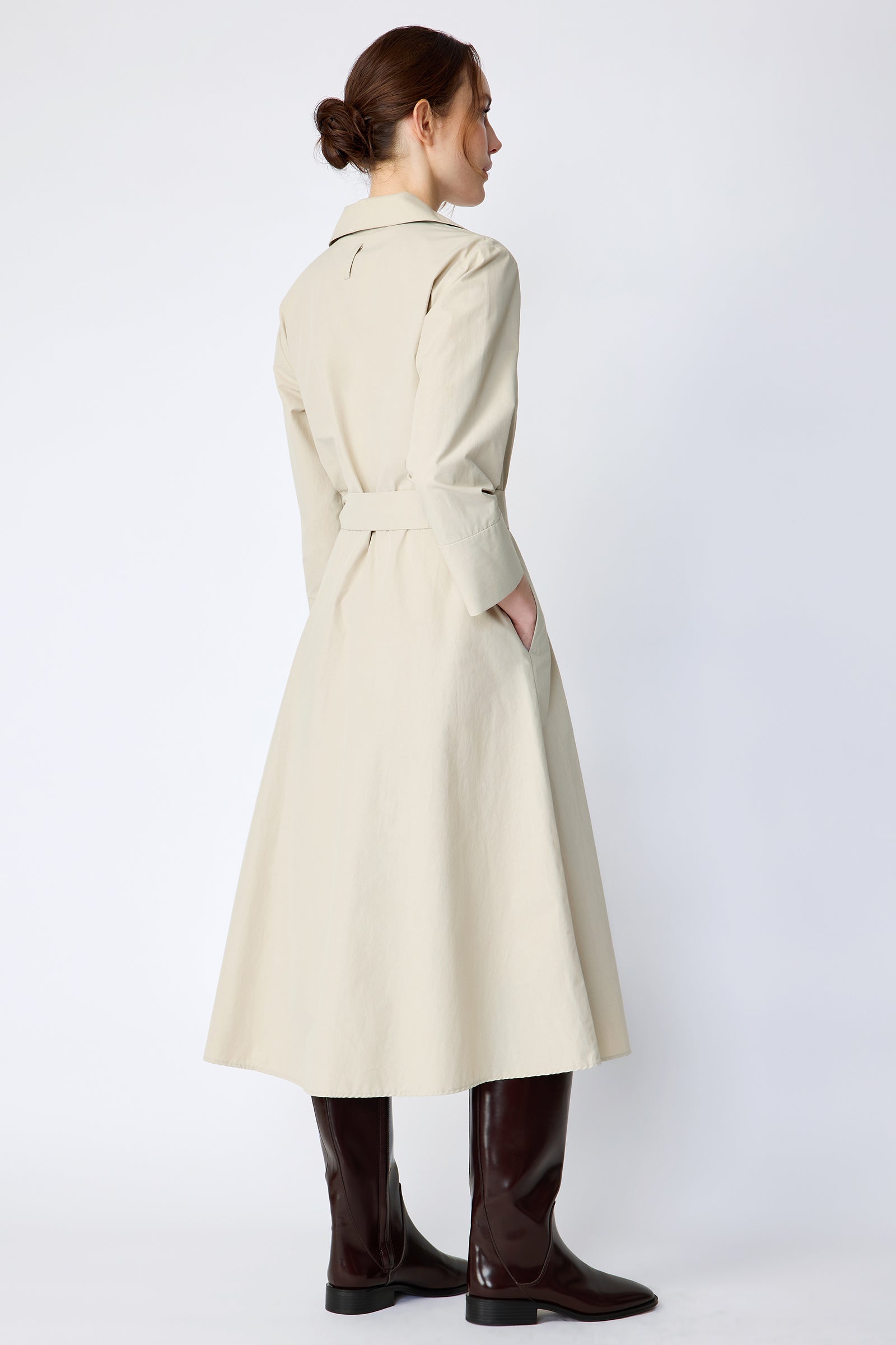 Woman wearing Kal Rieman Sydney Dress in Classic khaki on a white background with brown boots, hand in pocket, back view