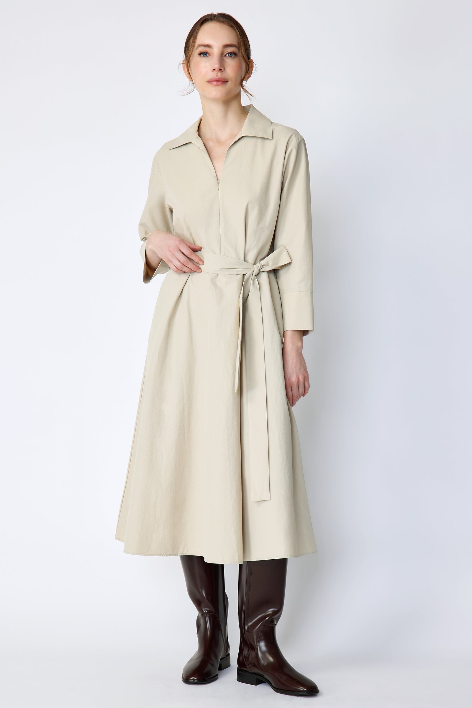 Woman wearing Kal Rieman Sydney Dress in Classic khaki on a white background with brown boots, hand on belt, arm on side