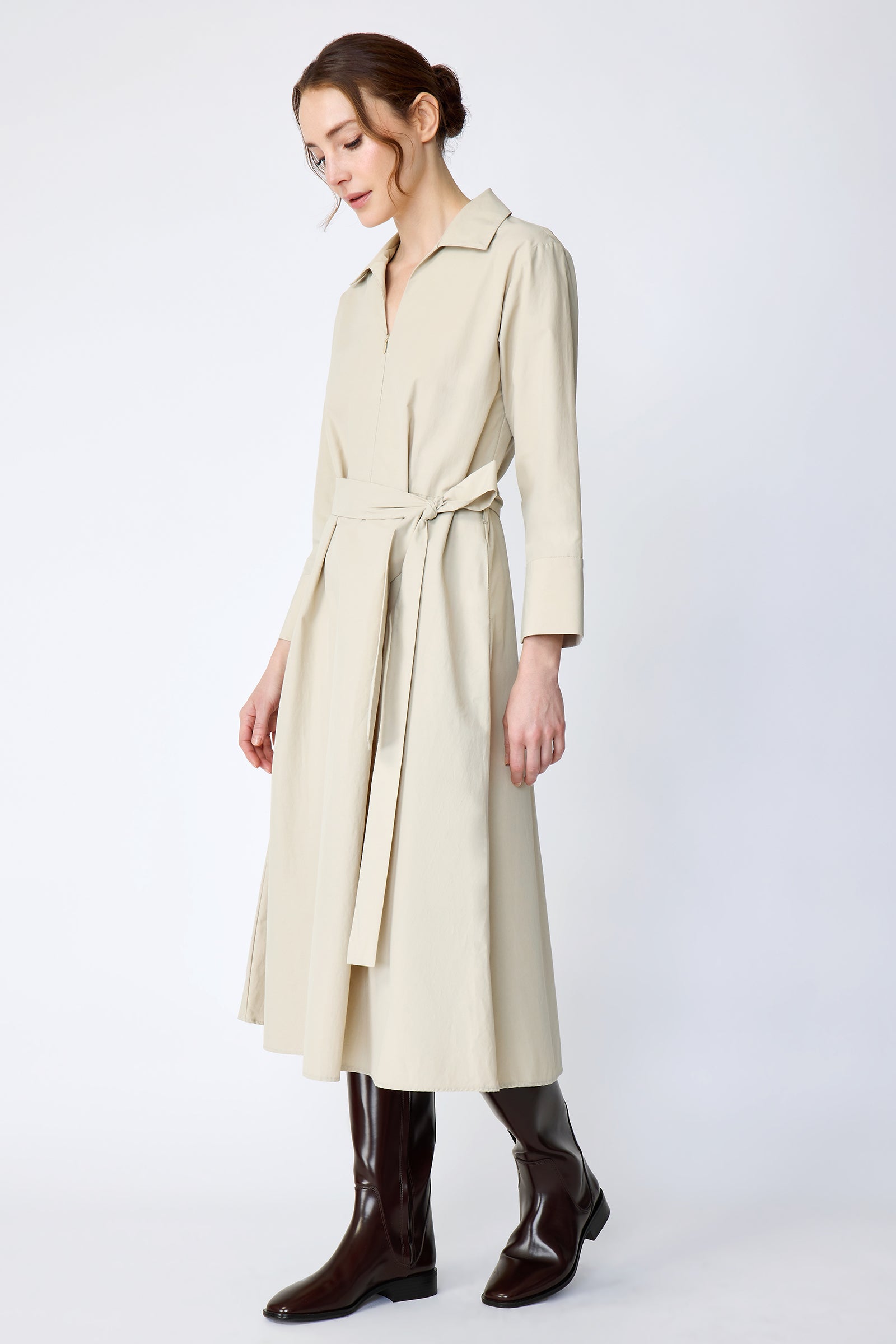 Woman wearing Kal Rieman Sydney Dress in Classic khaki on a white background with brown boots, arms on the sides, in movement, looking down