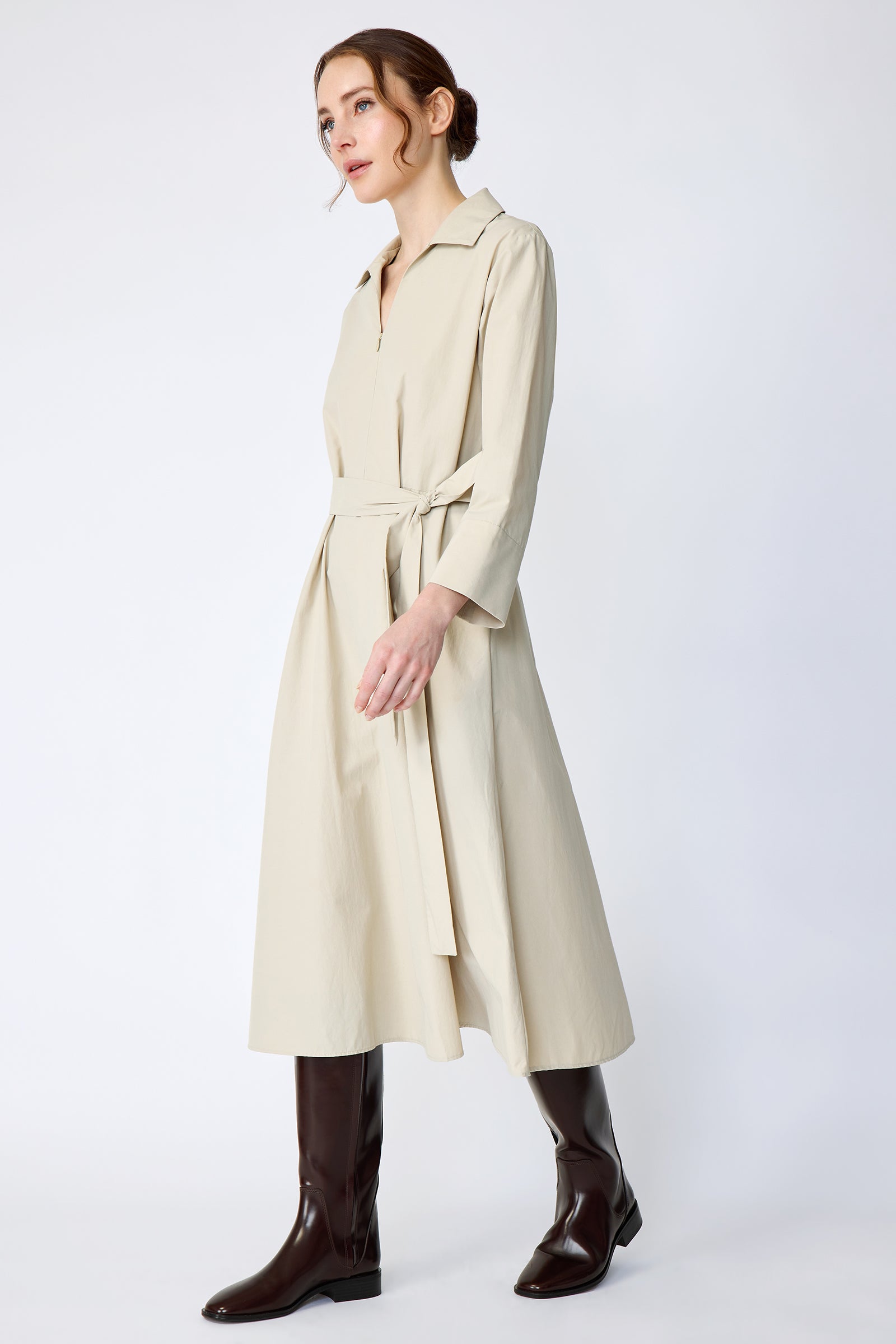 Woman wearing Kal Rieman Sydney Dress in Classic khaki on a white background with brown boots, arms on the sides, walking